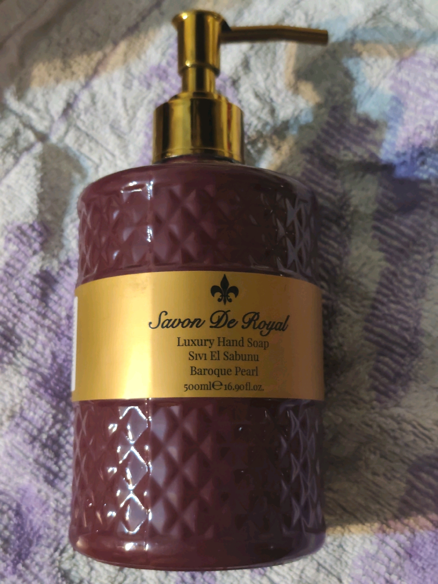 A close-up shot shows a dark red, textured bottle of hand soap. The bottle has a gold pump dispenser at the top and a gold label with black text. The text on the label reads "Savon De Royal," "Luxury Hand Soap," "Sivi El Sabunu," "Baroque Pearl," and "500ml 16.90 fl. oz." The bottle is placed on a purple and white textured surface, possibly a towel or blanket. The background is out of focus. The image does not provide any information about its location in a city.