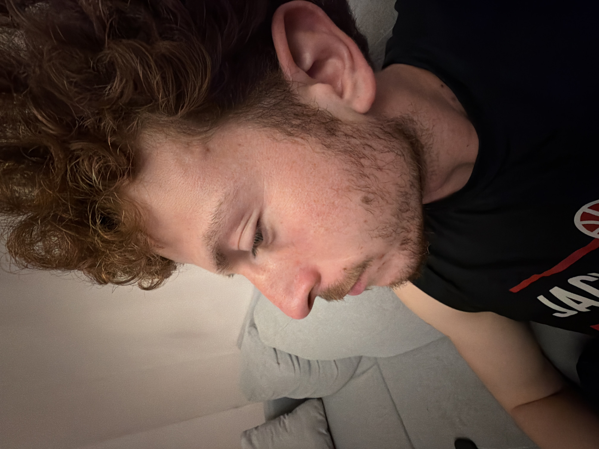 A close-up, rotated image shows a light-skinned person with reddish-brown curly hair and a short beard lying on a gray couch. The person is wearing a black t-shirt with a partially visible logo. The person's eyes are closed. The scene is dimly lit. No specific location in New York City is discernible from the image.