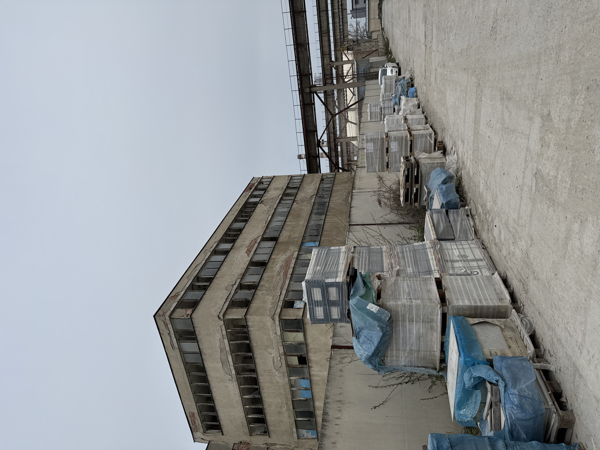 The image depicts an industrial site in Busmantsi, Bulgaria. On the left, a decaying, multi-story concrete building with numerous empty window frames rises into a gray sky. Adjacent to the building, a row of palletized materials wrapped in plastic stretch into the distance. Overhead, a network of metal pipes and support structures runs parallel to the stacked materials. A concrete ground surface is visible. No people are visible.