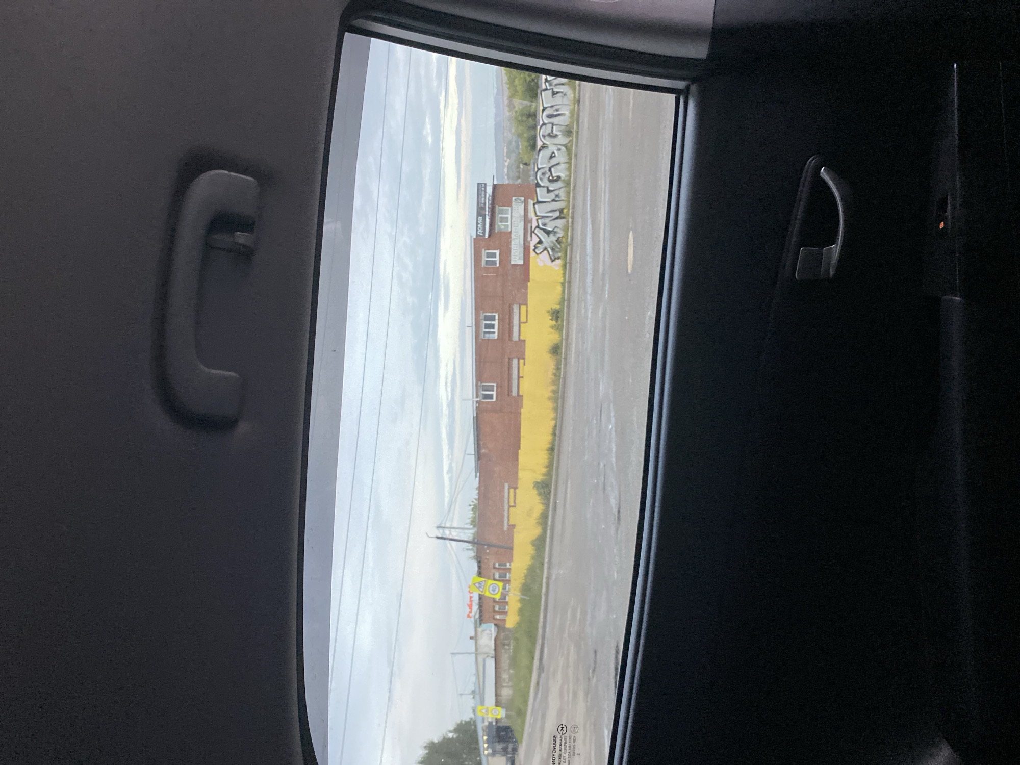 The image, taken from inside a vehicle, shows a view through the rear window. The scene outside is a street in Votkinsk, Russia, under an overcast sky. There are buildings with graffiti on them, one is red brick and another is yellow. A street runs in front of the buildings. There are a few traffic signs visible. The time of day appears to be daytime. The mood is neutral.