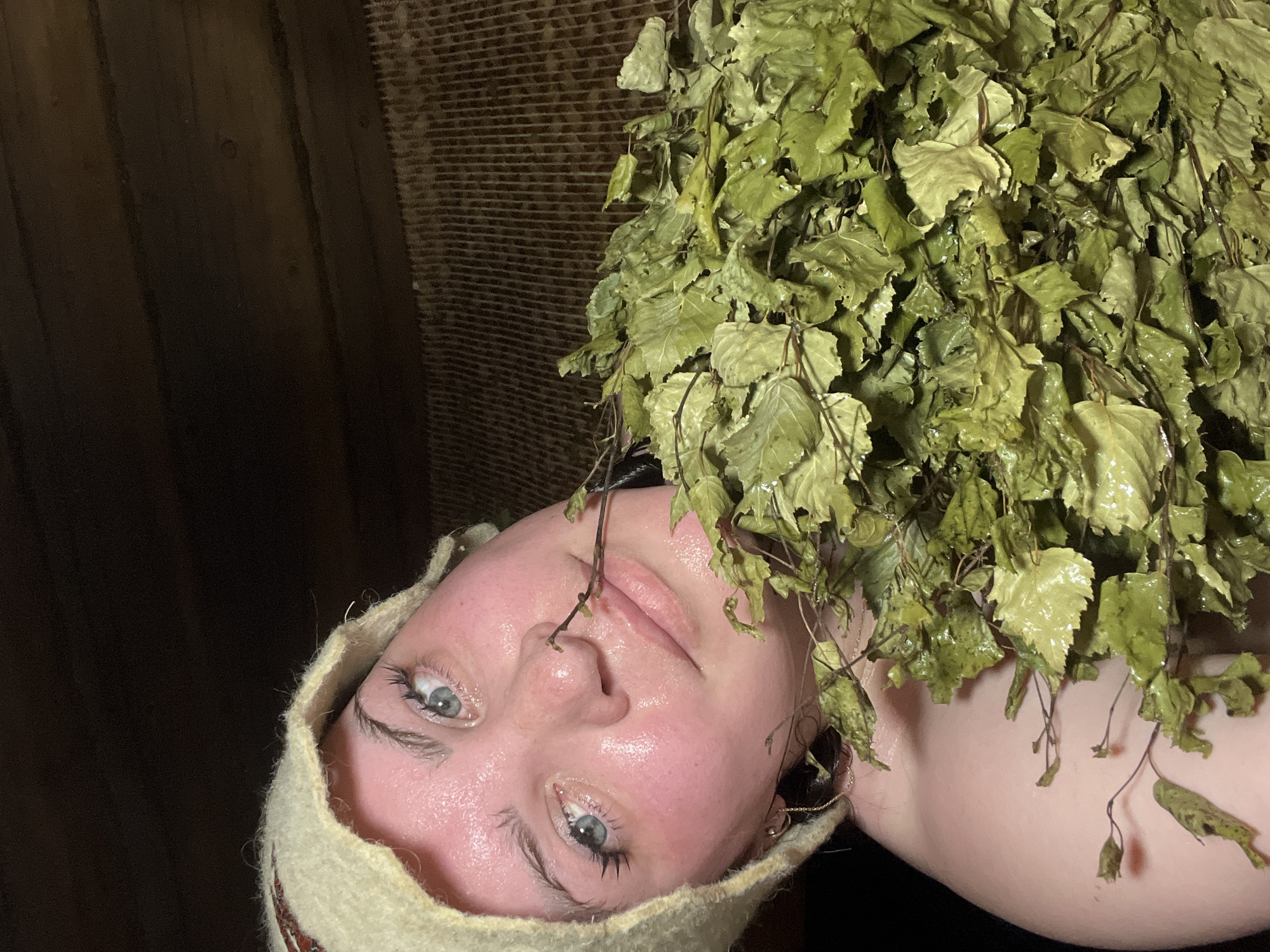 A person, presumably female, wearing a wool hat, is lying on their back, their face wet with perspiration or water. A bundle of leafy green branches, possibly birch, is positioned above their face, extending from the right side of the frame. The background features dark wood paneling on the left and a textured, woven wall covering on the upper right. The location is unspecified but presumed to be in Panteleevka, Russia.