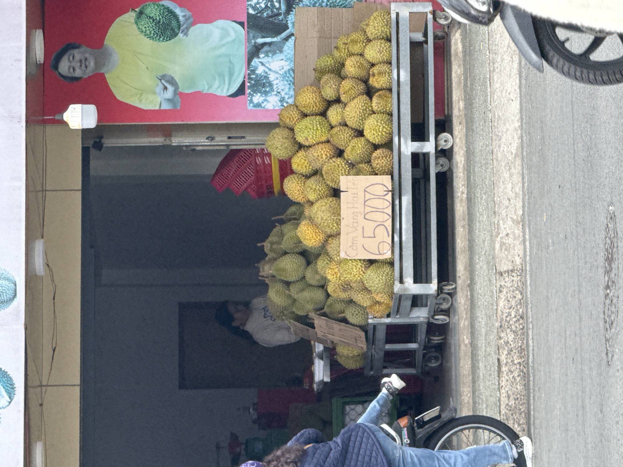 The image depicts a street scene in Da Lat, Vietnam, outside a fruit stand. A large pile of durians is displayed on a metal cart. A sign in front of the durians reads "65.000", likely indicating the price per kilogram. Behind the durians, a woman wearing a white sweater is visible inside the stand. To the right of the durians, a large poster features a man in a yellow shirt holding a durian. In the foreground, a person on a motorcycle, wearing a blue jacket and jeans, is positioned on the left side of the frame, with their left leg extended. Another motorcycle is partially visible on the right side of the frame. The ground in front of the fruit stand is paved.