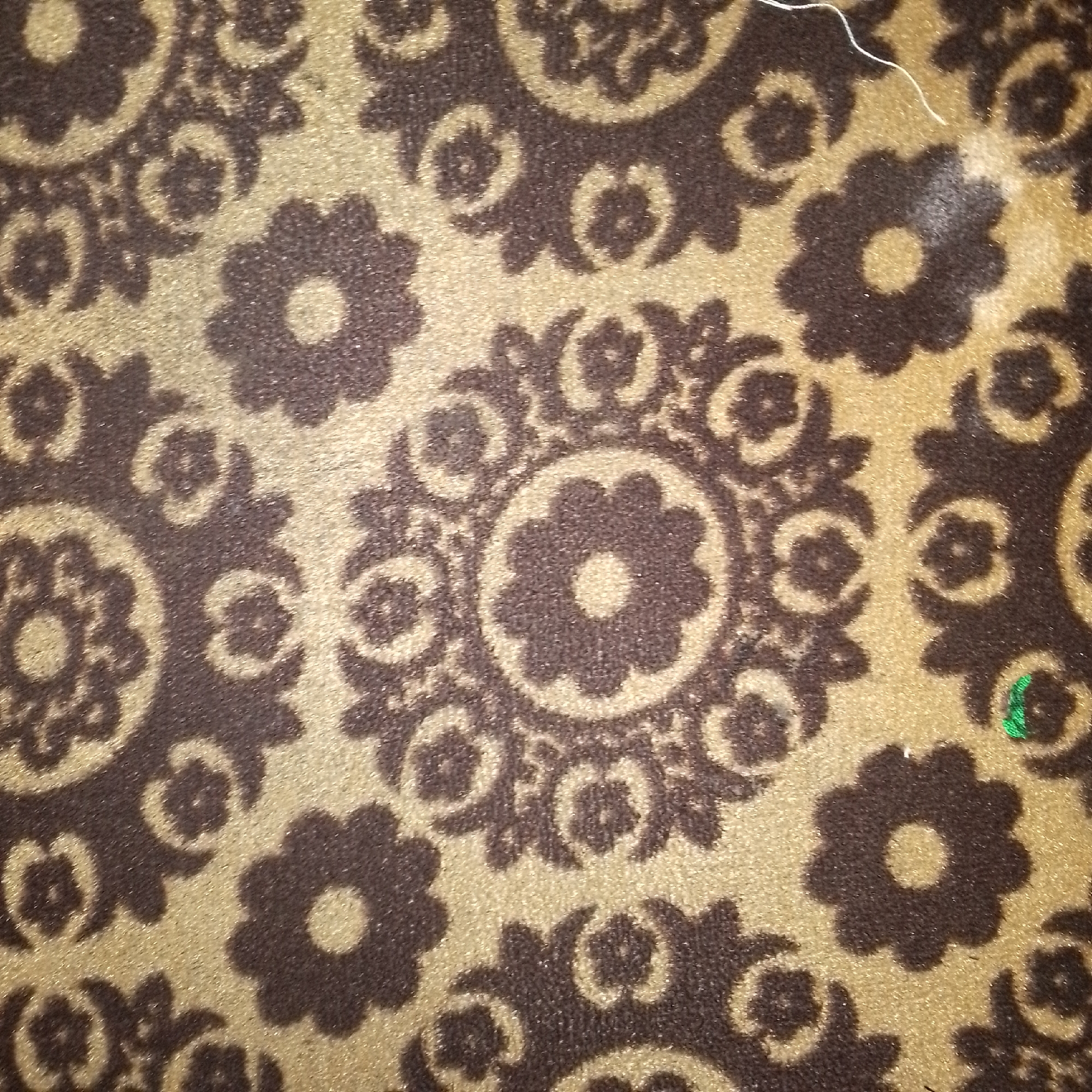 The image displays a close-up view of a textured surface with a repeating floral pattern. The pattern consists of dark brown, stylized flowers with lighter brown or golden centers, set against a textured, golden background. The flowers are arranged in a somewhat ornate, almost mandala-like fashion, with intricate swirling elements connecting them. The texture of the material appears to be fabric, possibly a carpet or upholstery, with a visible weave. There is a single strand of white thread running diagonally across the upper right portion of the image, and a small patch of green embroidery or thread is visible in the lower right quadrant. The lighting suggests it is an indoor setting, with a slight sheen on the golden material indicating a reflective quality. The image does not contain any identifiable landmarks or features that would allow for localization to Jauro Yinu, Nigeria, or any specific city.