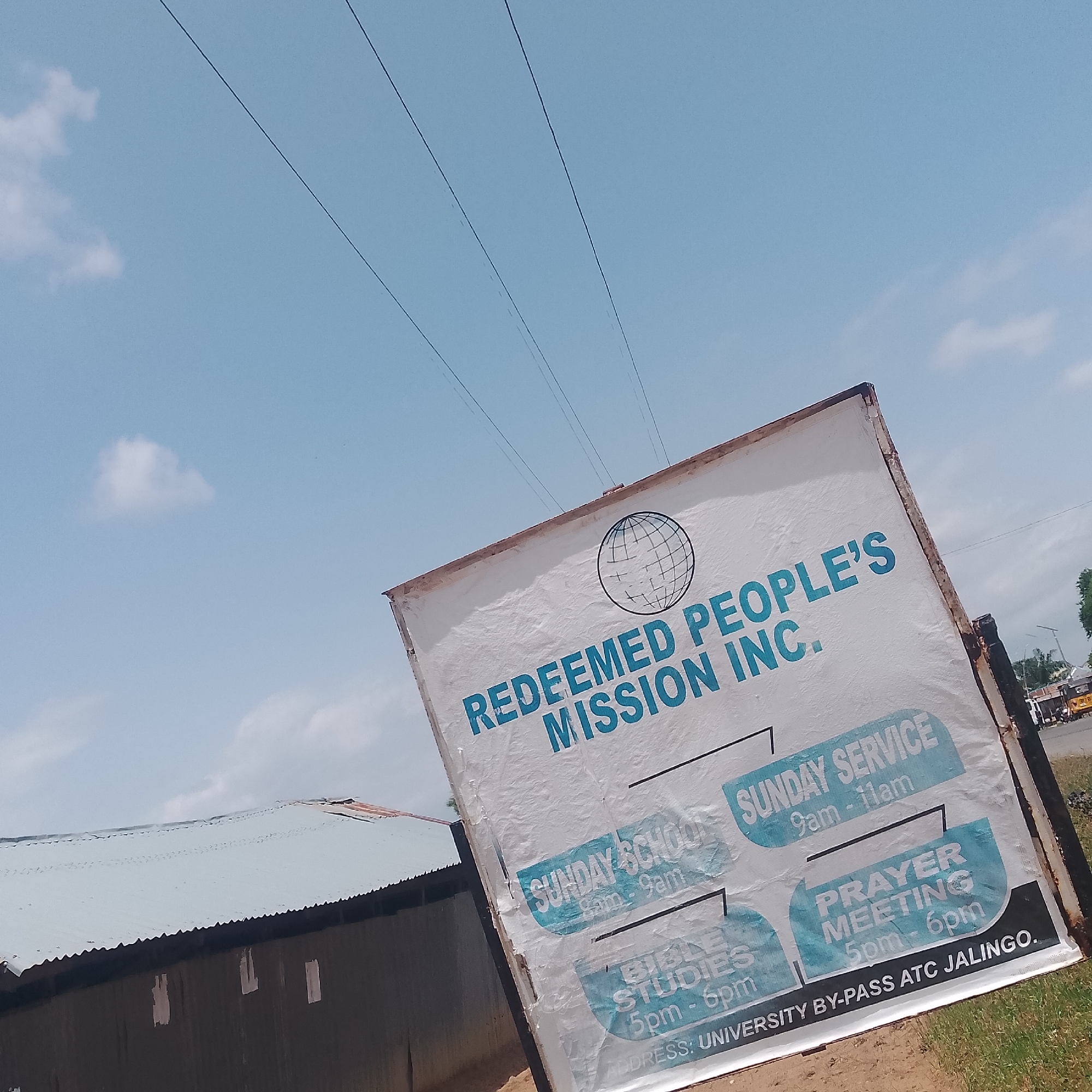 The image shows a weathered banner for "REDEEMED PEOPLE'S MISSION INC." The banner is tilted and positioned outdoors, with a clear blue sky and some scattered clouds visible. The banner displays information about church activities: "SUNDAY SCHOOL 9am - 9am", "SUNDAY SERVICE 9am - 11am", "BIBLE STUDIES 5pm - 6pm", and "PRAYER MEETING 5pm - 6pm". At the bottom, the address is listed as "UNIVERSITY BY-PASS ATC JALINGO." In the background, a corrugated metal roof of a building is partially visible to the left, and to the right, a road with a yellow vehicle and other structures can be seen, suggesting an urban or semi-urban environment. The overall scene depicts a roadside religious institution's signage.