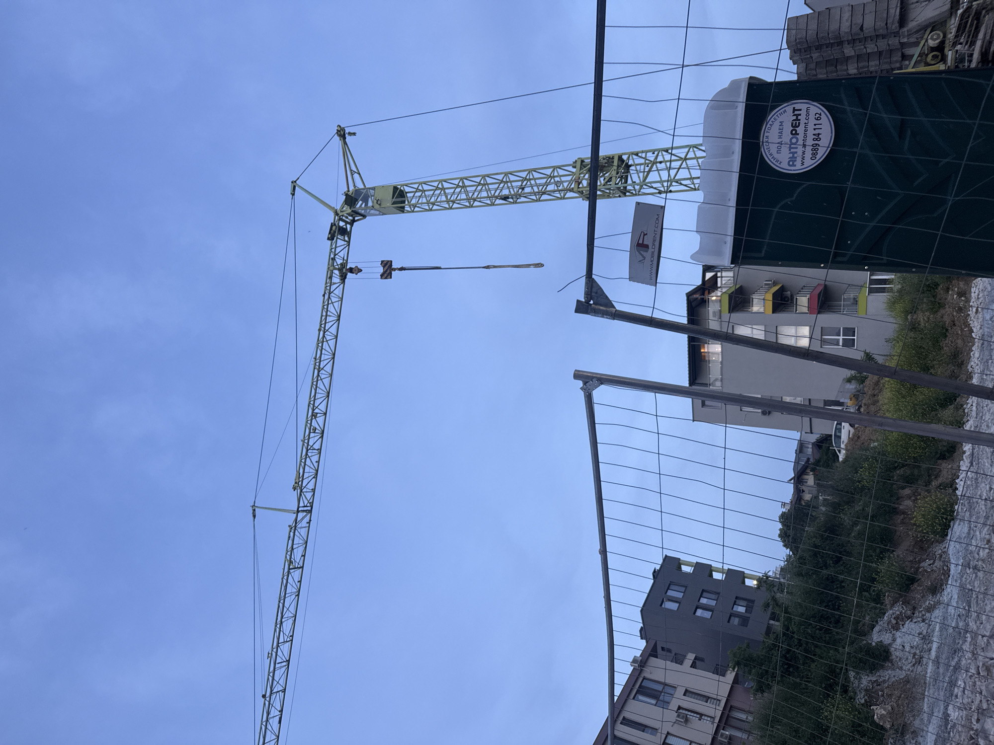 The image captures a construction site in Varna, Bulgaria, under a twilight sky. A large yellow construction crane dominates the upper portion of the frame, its boom extending horizontally across the sky. In the foreground, a temporary metal fence encloses the site. To the right of the fence, a green portable toilet is visible, featuring a white lid and a circular sticker with Bulgarian text and a phone number. Behind the fence, several multi-story buildings are under construction or have been recently completed, with balconies and windows visible. Lush green trees and bushes cover the sloped terrain between the buildings and the fence. The ground within the construction site is uneven, with piles of rocks and construction debris. The overall impression is one of active urban development.