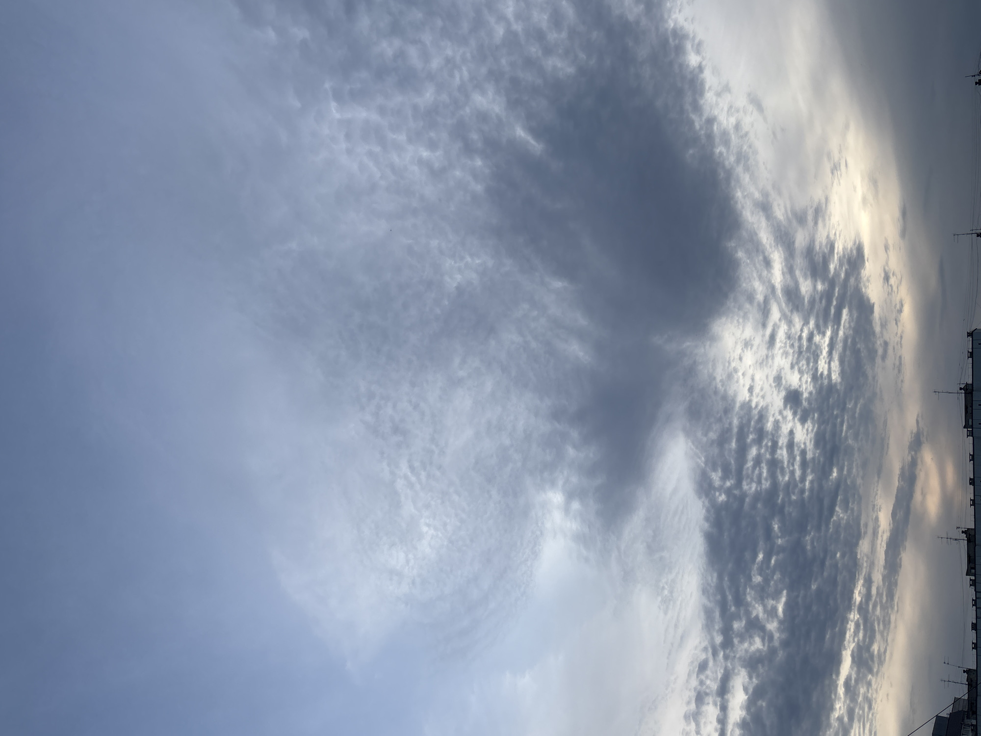 The image depicts a cloudy sky over the city of Chaikovsky, Russia. The clouds vary in density and texture, ranging from thin, wispy formations to denser, darker patches. Along the right edge of the image, buildings with antennas are visible.