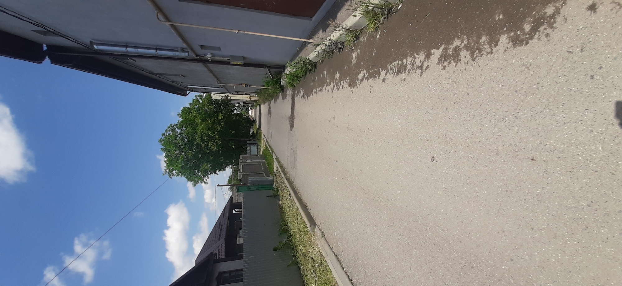 The image depicts a narrow paved street in the city of Yessentukskaya, Russia, on a bright, sunny day with scattered white clouds. On the left side of the street, there is a gray metal fence and a residential building with a dark roof. Further down the street, a large, lush green tree stands prominently. On the right side, a two-story building with a light gray facade and dark brown doors is visible. A vertical metal pipe runs down the side of this building, alongside a beige pipe. The street surface shows signs of moisture and some scattered debris. The scene appears to be a quiet residential area.