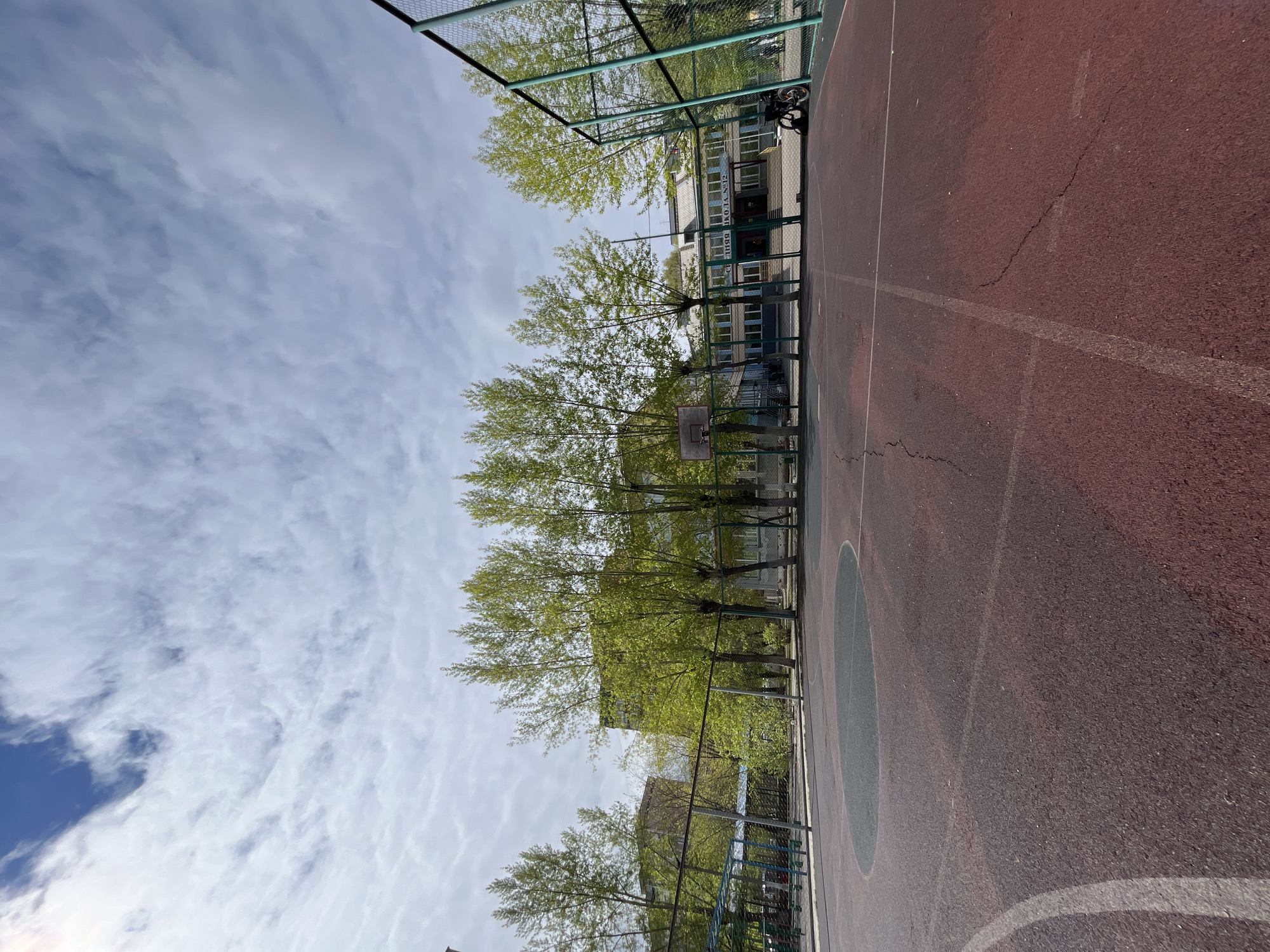 The image depicts a red and gray outdoor basketball court. A green fence surrounds the court, and trees with light green leaves are visible behind the fence. Beyond the trees, a building with windows and a sign in Russian ("С ОДОБРЯЮ ШКОЛА №2") can be seen. The sky above is filled with scattered white clouds. The location is identified as the city of Krasnoyarsk, Russia. A black object (possibly a bicycle) is leaning against the fence. The court shows signs of wear, including cracks in the surface. White lines mark the boundaries and key areas of the basketball court.