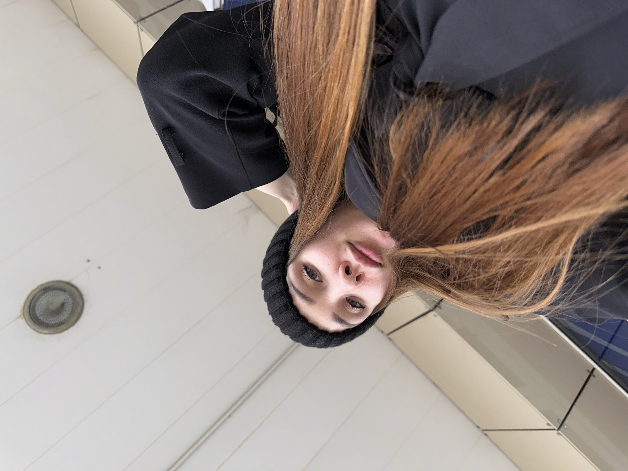 A young woman with long auburn hair is looking down at the camera. She is wearing a black beanie and a dark-colored coat or jacket. The background is a white tiled wall with a circular vent visible in the upper left corner. There is also a metallic or mirrored section visible in the background. The image has a distorted perspective, as if the camera is directly below the subject. The location is presumed to be Chaikovsky, Russia.