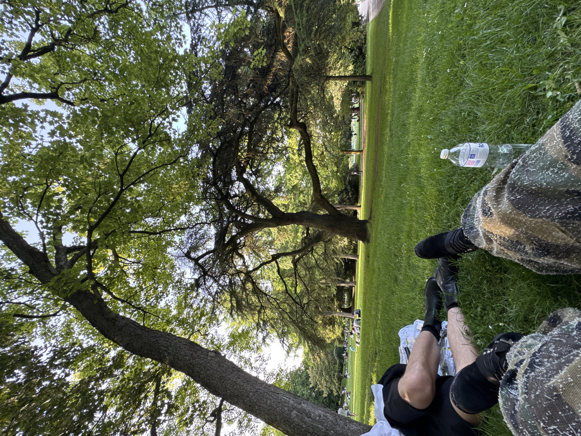 The image depicts a scene in a grassy area, likely a park or cemetery, with multiple trees overhead providing partial shade. In the foreground, a person is sitting on the grass, wearing black shorts, black boots, and a camouflage patterned garment covering their lap. A half-empty plastic water bottle lies on the grass near them. In the distance, other individuals are visible sitting or lying on the grass. The location is likely a park or cemetery, but I have no way to determine that the location is in the city of Paris, France.