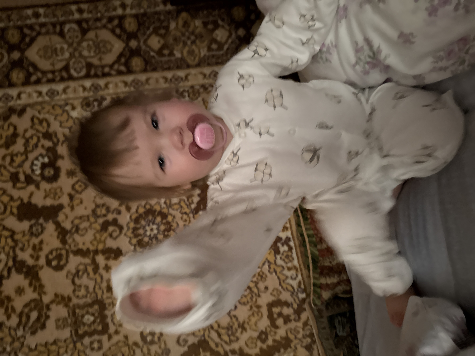 A baby with a pacifier is lying on a rug. The baby is wearing a patterned onesie. The rug has a dark brown and tan pattern. The baby is partially covered by a blanket with a floral pattern. The scene appears to be indoors. No location information for Krapivka, Russia, can be identified from the image.