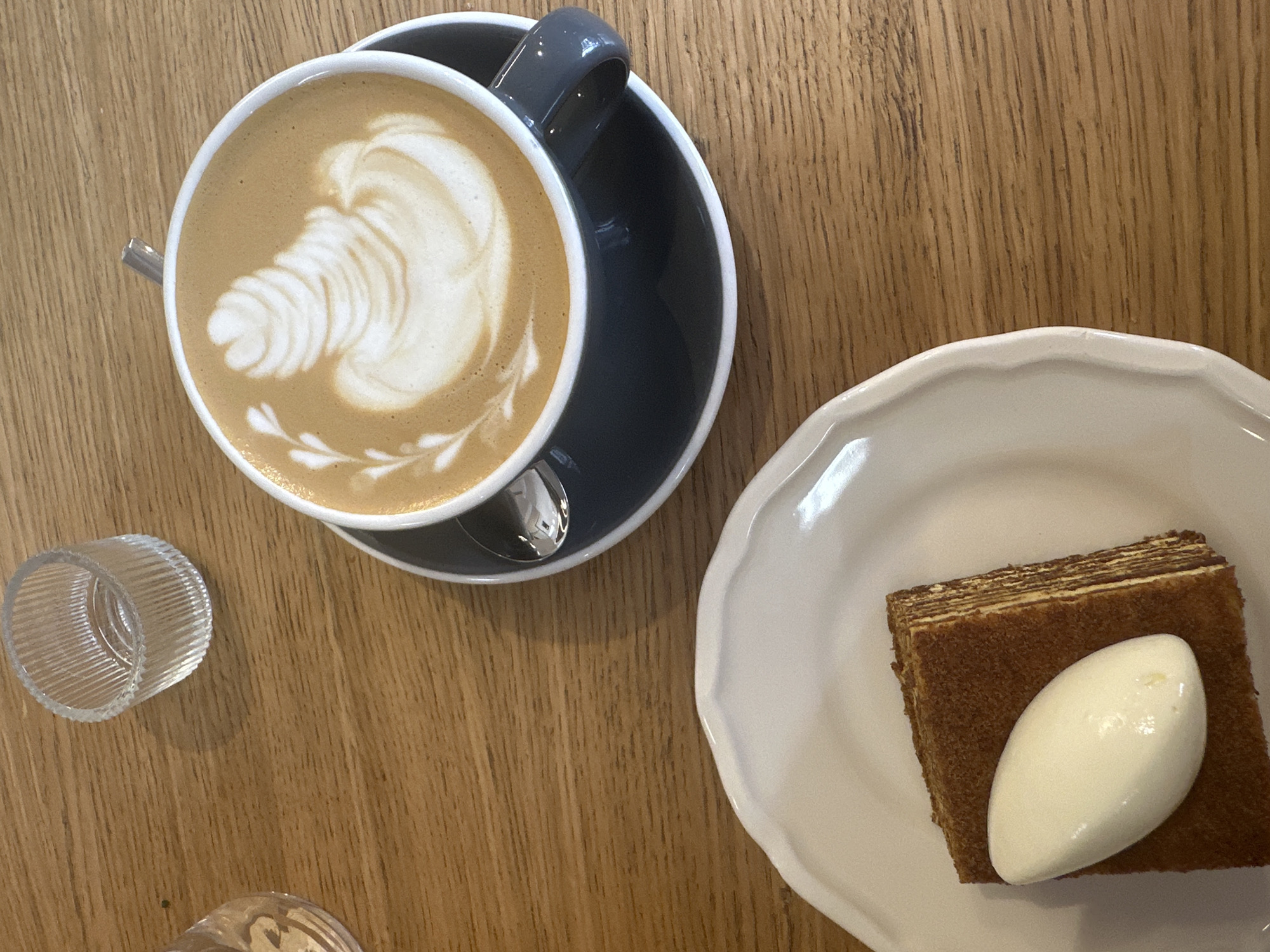 The image shows a close-up, overhead view of a wooden table in a cafe, likely in Vracar Urban Municipality, Serbia. On the table, there is a dark grey saucer holding a white cup of coffee with latte art. To the left of the coffee cup, a small, clear, ribbed glass container is visible. In the foreground, a white plate holds a slice of layered cake topped with a quenelle of light-colored cream or ice cream. The overall scene depicts a moment of enjoyment of coffee and dessert.
