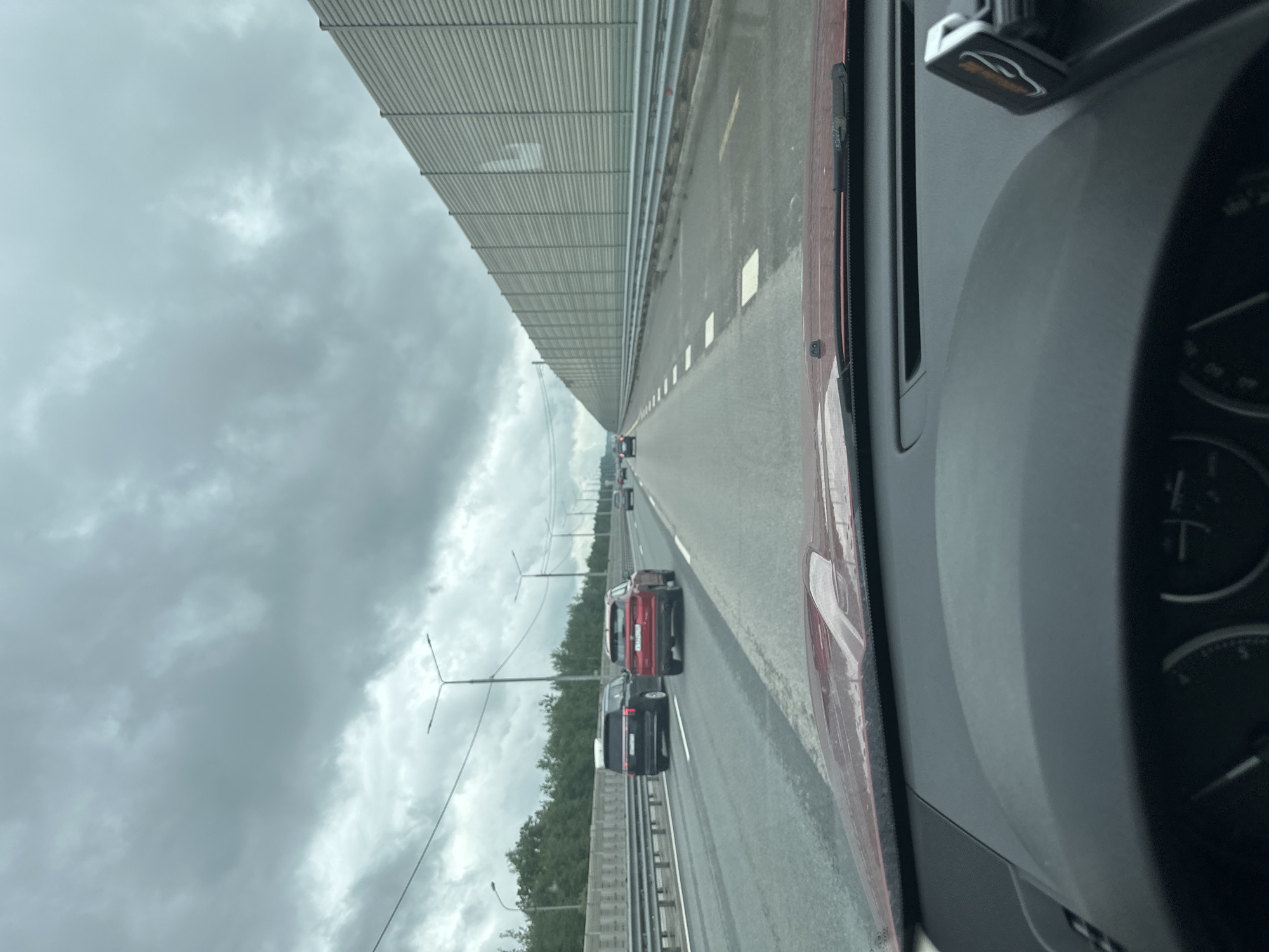 The image is a forward-facing view from inside a vehicle, likely in motion on a highway. The sky is overcast with dark, heavy clouds. A large, grey, corrugated sound barrier stretches along the right side of the road, extending into the distance. On the road, there are several cars. In the left lane, a black SUV is visible, followed by a red SUV in the right lane. Further ahead, other vehicles are present in both lanes. The road surface appears to be asphalt, marked with white lines and squares. Streetlights are visible on poles on the left side of the highway, above a safety barrier and trees. The dashboard of the car is partially visible in the foreground, with part of the windshield also in view. The overall impression is a mundane scene of highway travel under cloudy skies.