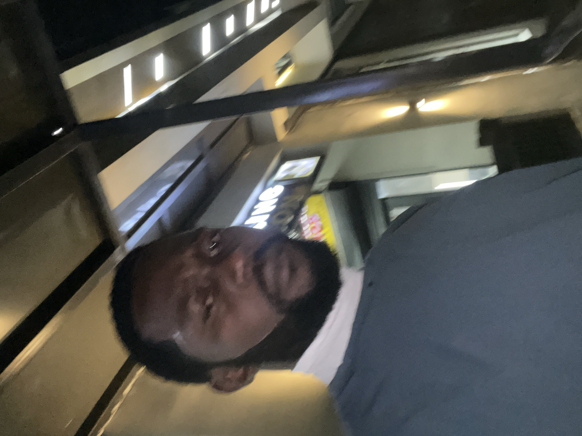 The image is a slightly blurry, low-angle, outdoor selfie taken at night in Guzape, Nigeria. The subject is a Black man with a full beard and short, dark hair, wearing a dark blue long-sleeved shirt over a white turtleneck. He is looking directly at the camera with a serious expression. In the background, a building with a lit sign is visible, featuring illuminated white lettering. The environment appears to be an urban setting with architectural elements and lighting suggesting a commercial area.