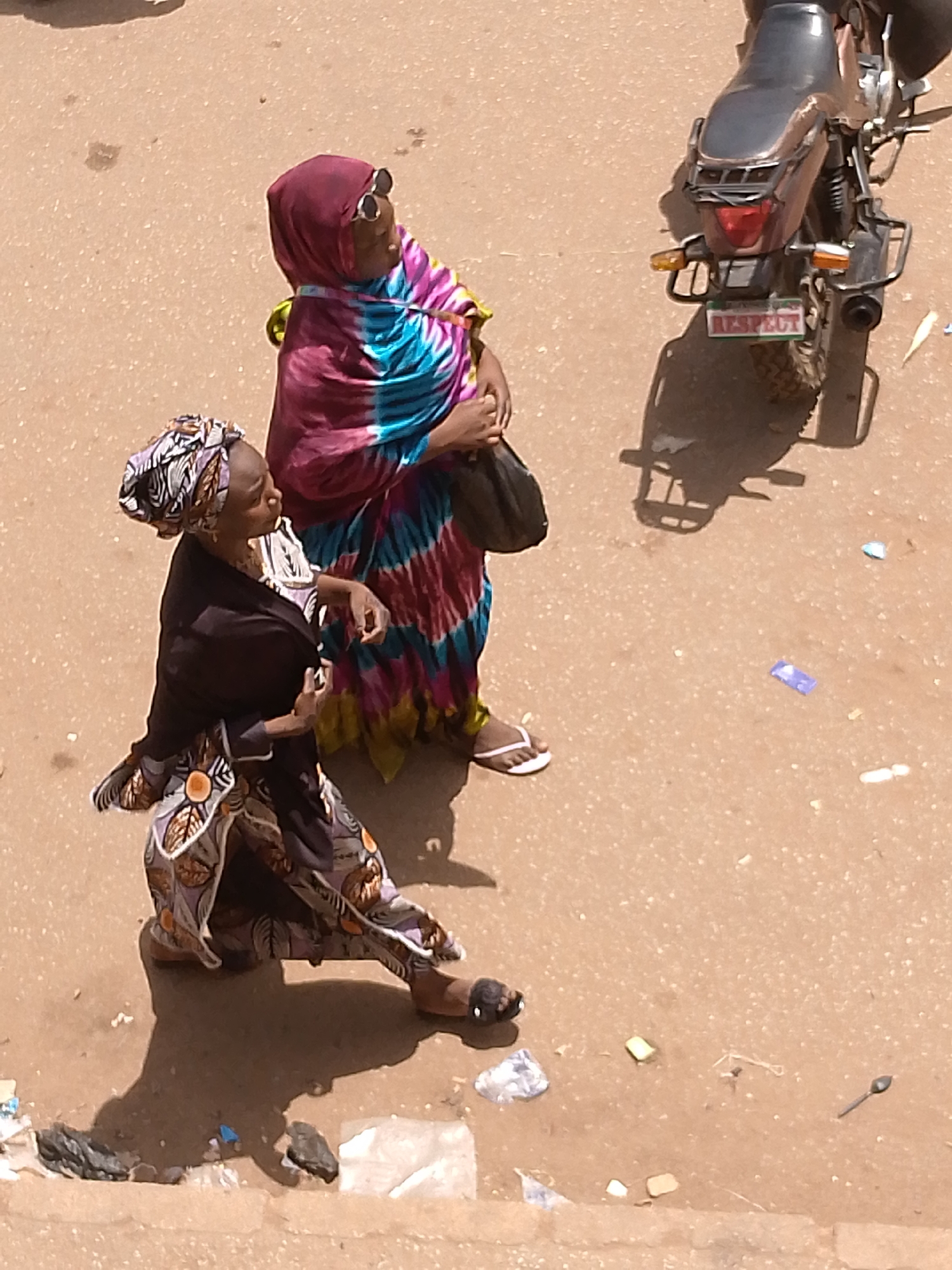 A high-angle outdoor shot depicts two women and a motorcycle on a paved surface in Bukuru, Nigeria. The woman on the left wears a dark jacket over a patterned dress and headwrap. The woman on the right wears a pink headscarf, sunglasses, and a colorful tie-dye-like dress, carrying a black bag. A motorcycle, oriented vertically, is positioned behind the women. Litter is scattered on the ground. The motorcycle's license plate reads "RESPECT".