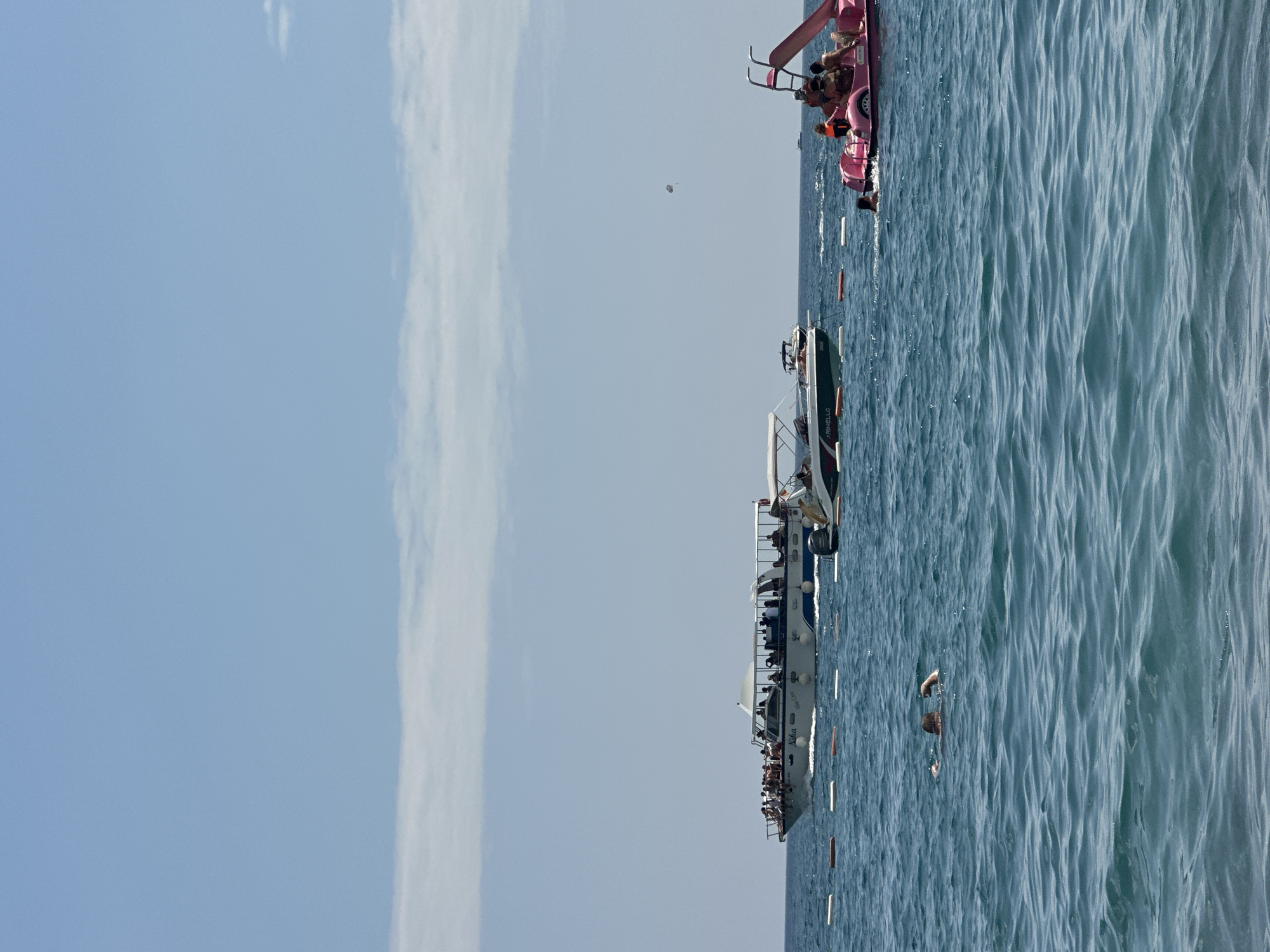In Boreti, Montenegro, on a sunny day, several boats are in the turquoise water. A large white boat with a blue stripe is filled with people. A smaller white boat with an awning is also present. To the right, a pink paddleboat with a slide carries several people, including children wearing life jackets. In the foreground, a person is swimming in the water. The sky is blue with scattered white clouds.
