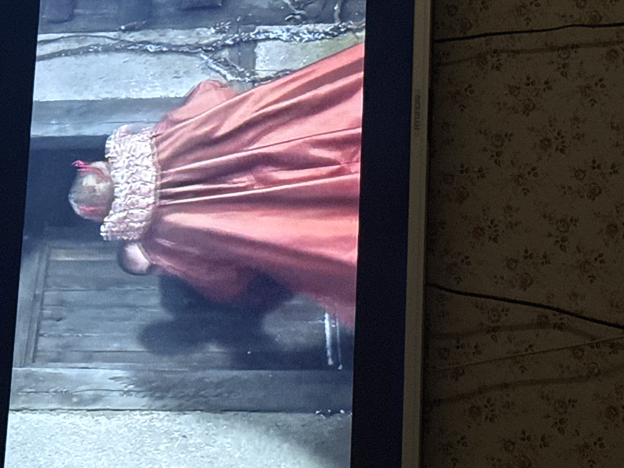 The image shows a television screen displaying a scene of a person in a red gown lying on a wooden platform or stairs. The person is positioned head-down with their head facing the top of the screen. Their fair hair is visible. The gown has a ruffled neckline. The platform or stairs appears to be made of rough-hewn wood and is set against a background that looks like stone. The word "HYUNDAI" is partially visible along the right edge of the screen. Adjacent to the screen is a floral patterned surface. The image does not provide any information to confirm or deny the location of the image in Скалба Снт, Russia.