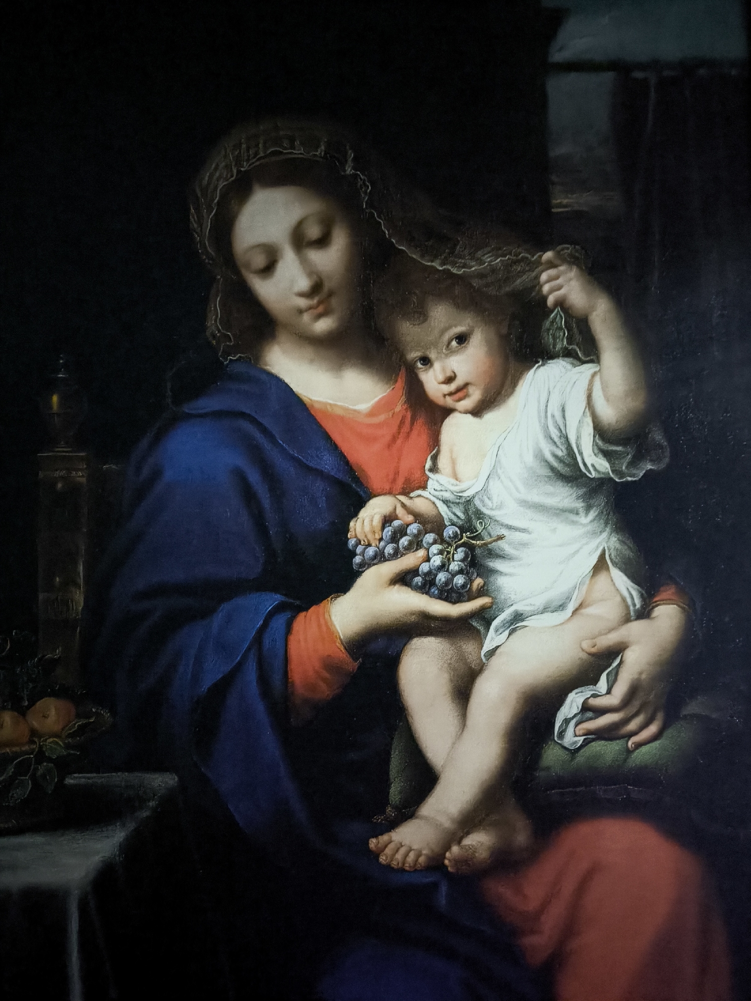 This is a painting depicting the Virgin Mary holding the infant Jesus. The Virgin Mary is wearing a blue mantle over a red garment and a veil on her head. She is looking down at the infant with a gentle expression. The infant Jesus is dressed in a white tunic and is reaching for the grapes held by the Virgin Mary. He is looking directly at the viewer with a curious expression. In the background, a dark curtain hangs on the right side of the painting, and on the left, a table holds a basket of fruit and a candlestick. The overall lighting is dim, creating a somber and intimate atmosphere. This painting is located in the Municipal Gallery of Kallithea, Greece.