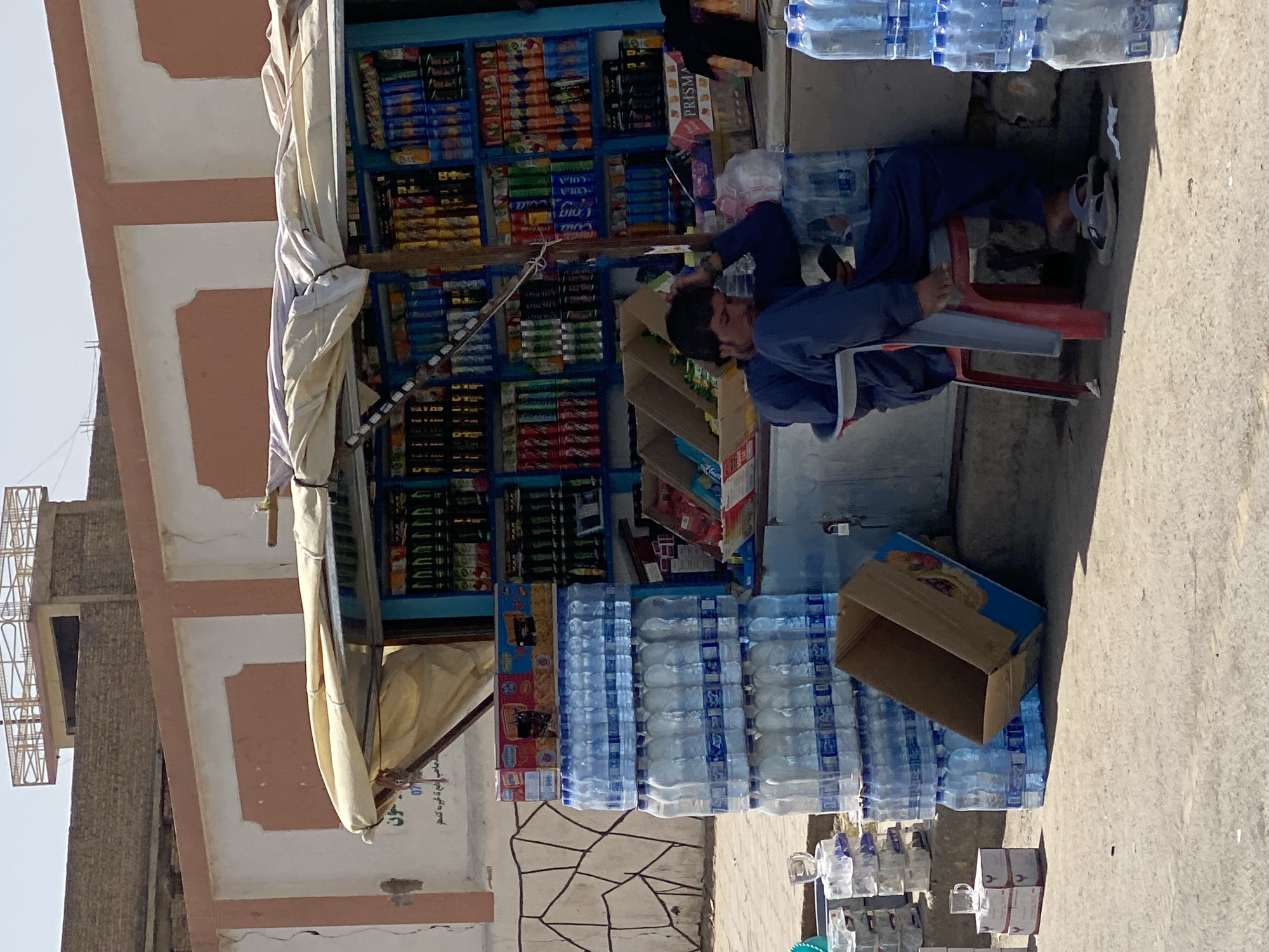 A man wearing a blue garment and a head covering sits on a red plastic chair in front of a small shop in Kandahar, Afghanistan. He is looking at his mobile phone. The shop is stocked with various products including bottled water, sodas, and snacks. Several stacks of bottled water are visible outside the shop. The shop is set against a building with a light orange and white facade. The ground is a dusty, concrete surface. The scene appears to be a street-level market or small store.