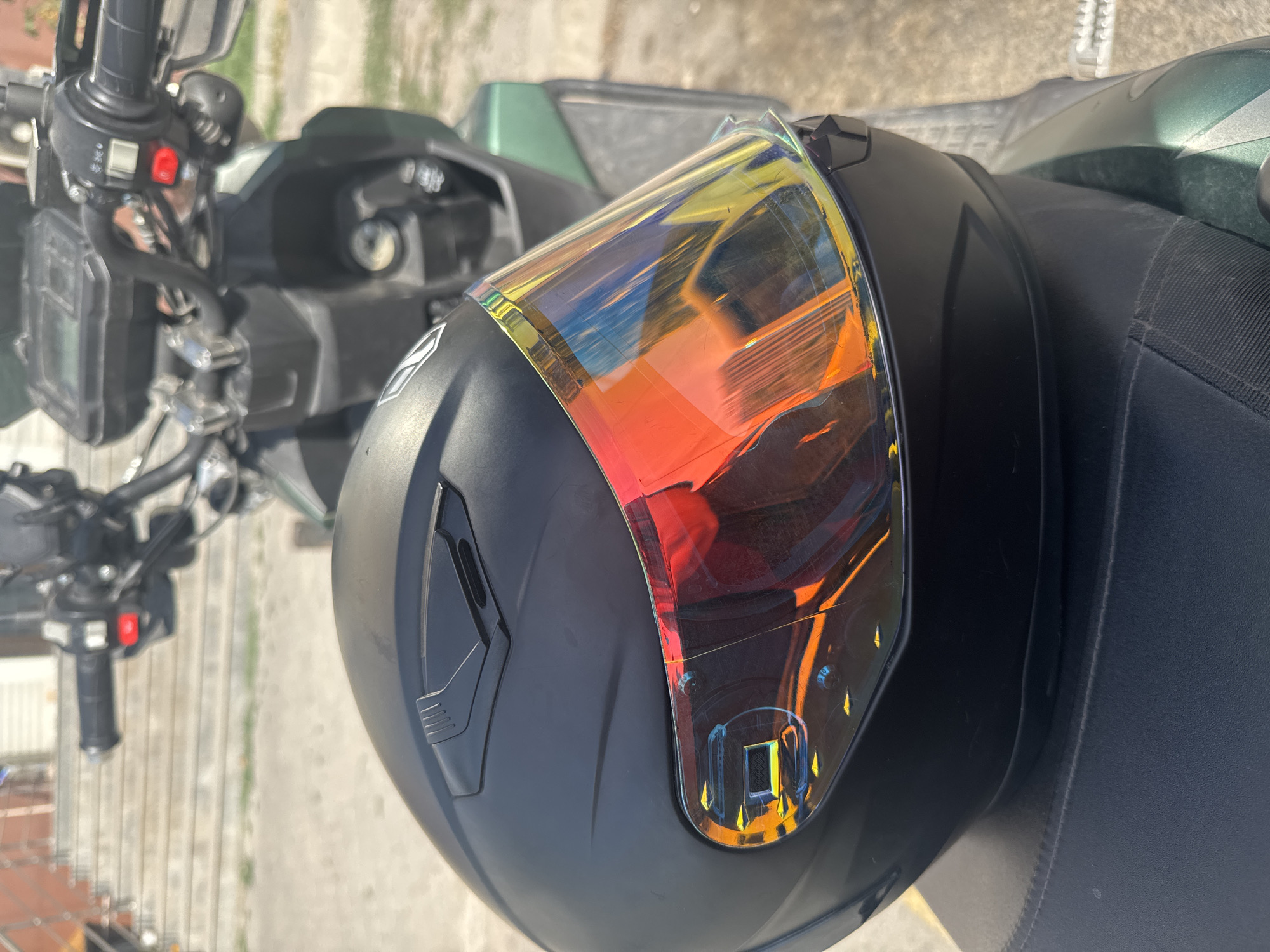 A matte black motorcycle helmet with a mirrored, iridescent visor sits on the seat of a green motorcycle. The motorcycle's handlebars and dashboard are visible in the background. The scene is outdoors, with a paved surface and some stairs in the distance. The lighting suggests it is daytime. The image appears to have been taken in Bishkek City, Kyrgyzstan, given the context of the prompt.