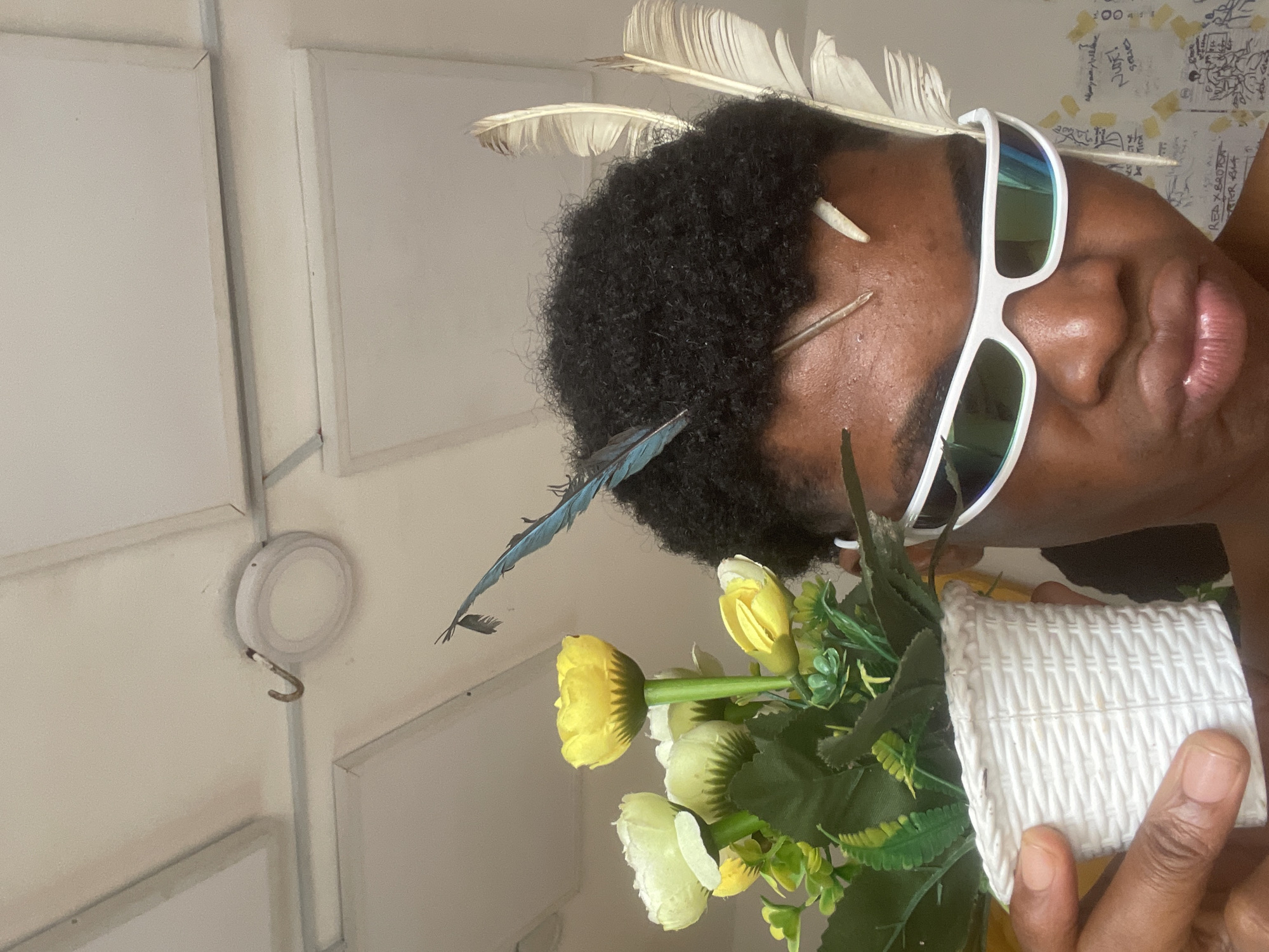 A person with dark, curly hair and white, futuristic sunglasses is holding a small white woven pot containing artificial yellow and white flowers. Feathers, including one blue and several light brown, are stuck in the person's hair. The background appears to be an indoor ceiling with white tiles and a light fixture. A hook is visible on the ceiling. The image is cropped to focus on the person's face and the flowers. The location is stated to be Ado, Nigeria, but there are no specific visual cues in the image to confirm this.