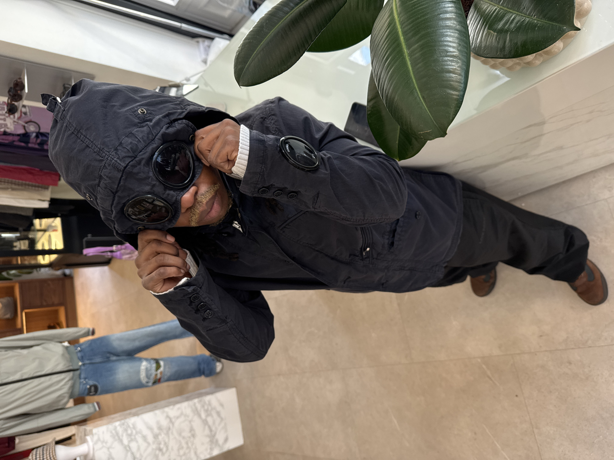 The image features a man indoors, likely within a retail store or boutique setting in London, UK. He stands centrally, looking directly at the viewer. The man, appearing middle-aged with a mustache and goatee, is wearing a distinctive dark blue or black hooded jacket. The hood incorporates two prominent, round, dark lenses or goggles, which are positioned over his eyes. His hands are raised, seemingly adjusting the hood or the lenses. A white ribbed cuff is visible at his left wrist, and a circular black patch with white text reading "C.P. Company" is visible on the jacket's left sleeve, near the cuff.
To the man's right, a large plant with glossy dark green leaves, resembling a rubber plant, rests on a pristine white surface, likely a counter or display table. The background to his left shows additional elements of a retail environment, including shelves displaying various items and, further back, a mannequin dressed in a grey jacket and light blue jeans. The floor throughout the space is covered with light-colored, possibly beige or cream, tiles. The overall impression is one of a modern, well-lit interior. The man's partial expression suggests a casual or playful demeanor.