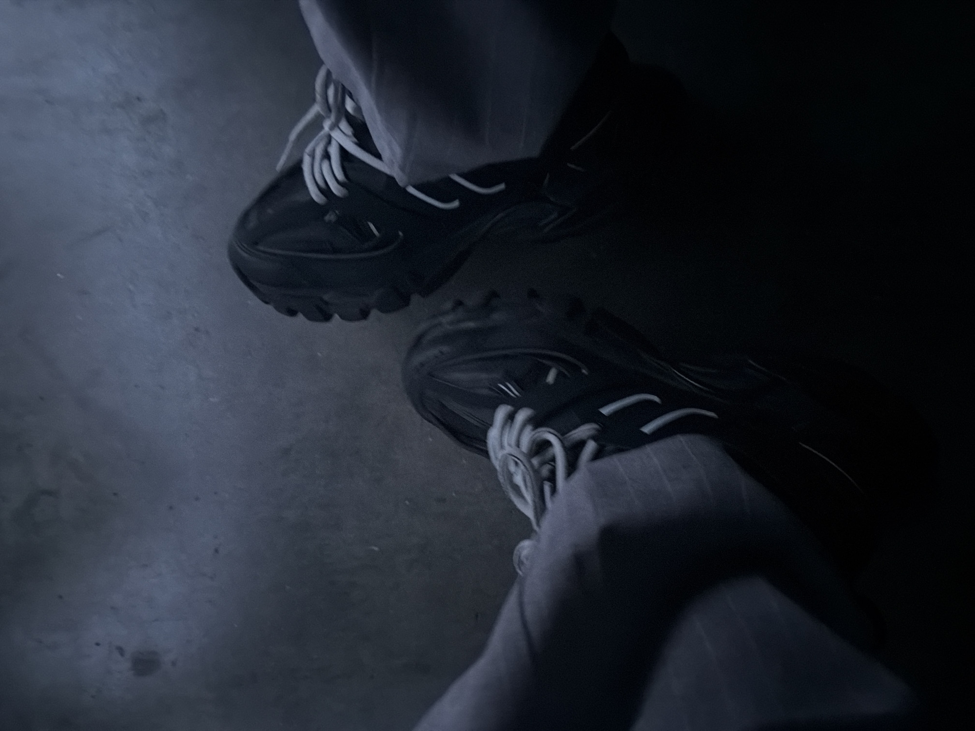 The image depicts a close-up, overhead view of a person's legs and feet, wearing dark-colored sneakers with white laces and grey pinstripe pants. The perspective suggests the person is either sitting or standing on a dark, textured surface, possibly concrete, which appears slightly damp or weathered. The lighting is dim, casting deep shadows and obscuring fine details of the environment. No specific landmarks or features of Zlatograd, Bulgaria, are visible in the frame, making it impossible to ascertain the location within the city based solely on the image content. The scene is static, with no apparent action or interaction taking place.