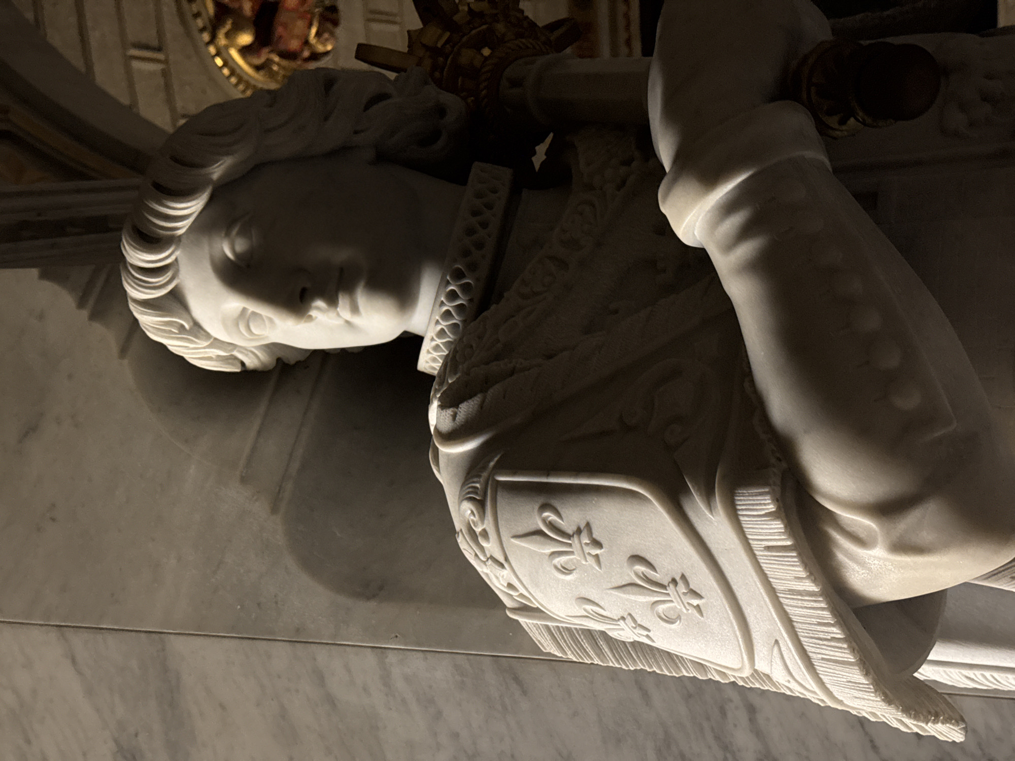 The image shows a marble statue of a male figure, likely a king or a saint, dressed in ornate armor. The statue is positioned indoors, with marble walls and a hint of golden architectural decoration in the background. The figure is depicted from the chest up, with a regal expression and elaborate detailing on the armor, including fleur-de-lis symbols on a shield. The statue is lit by a spotlight from the left, casting shadows that emphasize the texture and form of the marble. This statue is located in the Royal Pantheon of El Escorial Monastery, in San Lorenzo de El Escorial, Spain, specifically within the Basilica of the Monastery, which houses the tombs of Spanish royalty.
