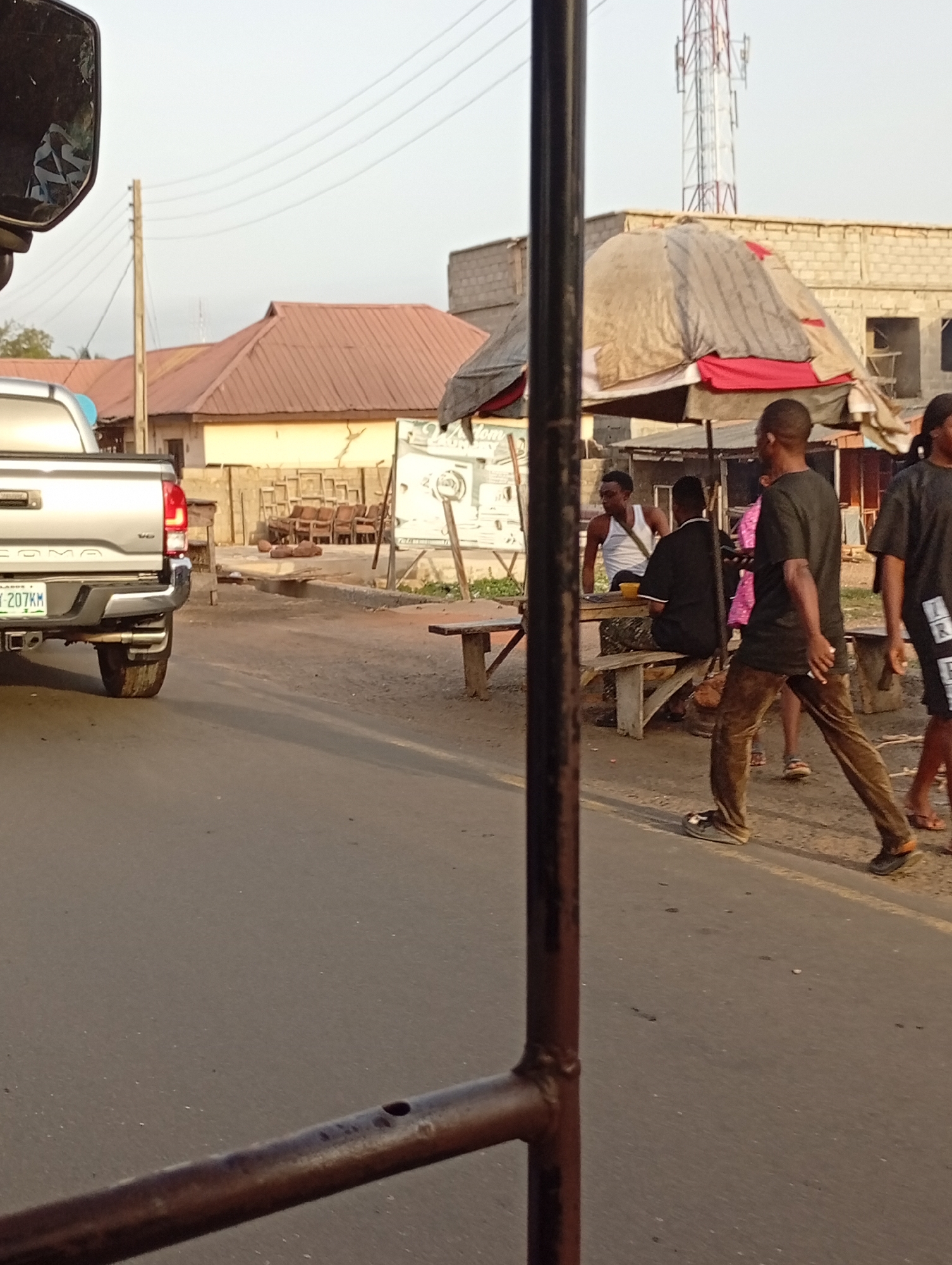 An outdoor scene captures a street or roadside in what appears to be a vibrant community. The photograph is taken from inside a vehicle, with its side mirror visible in the upper left corner and a dark metal pole vertically bisecting the frame.
In the mid-ground, a silver pickup truck, identifiable as a Toyota Tacoma by the partially visible "COMA" on its tailgate, is parked or moving slowly on the left side of the road. Its license plate clearly displays "LAGOS" in the top left corner and "Y 207KM" as its main identifier, indicating a location in Lagos, Nigeria.
On the right side of the road, several people are gathered around a makeshift stall or sitting area. A man in a white tank top is seated on a wooden bench, engaged with others, including another person seated with their back to the camera. Adjacent to them is a vendor's stall covered with a gray and red tarp. Two men are walking towards the right: one in a dark t-shirt and brown pants, holding a light-colored object, and another in a dark shirt and shorts.
The background features residential or commercial buildings with reddish-brown corrugated metal roofs and some unpainted concrete block structures. A prominent telecommunications tower rises in the distance. Wooden power poles and lines crisscross the bright, clear sky. Stacks of wooden furniture, possibly chairs and frames, are visible behind the truck.
The soft, warm lighting suggests either early morning or late afternoon. The overall atmosphere appears casual and everyday, with people going about their routines in a lively street setting.