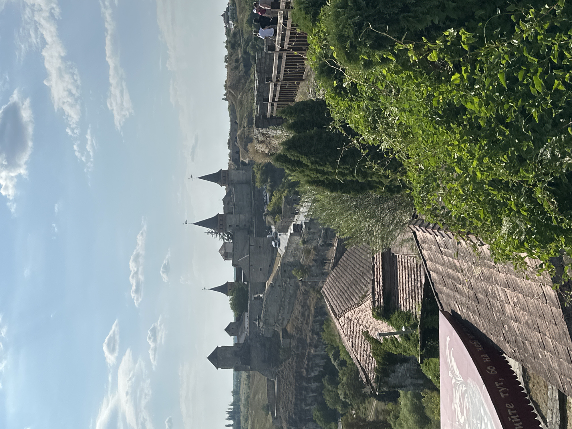 The image displays a panoramic view of the Kamianets-Podilskyi Castle in Ukraine. In the foreground, a tiled roof structure with a red awning bearing Cyrillic text is visible. Beyond this, a wooden walkway with a railing leads the eye towards the castle complex. Several people, including men and women, are seen walking along this walkway. The castle itself, a formidable stone structure with multiple towers and battlements, is perched atop a rugged cliff. A road with a few vehicles, including a bus and a car, runs adjacent to the castle walls. Lush green vegetation, including trees and bushes, surrounds the scene, with rolling hills and a partly cloudy blue sky forming the backdrop.