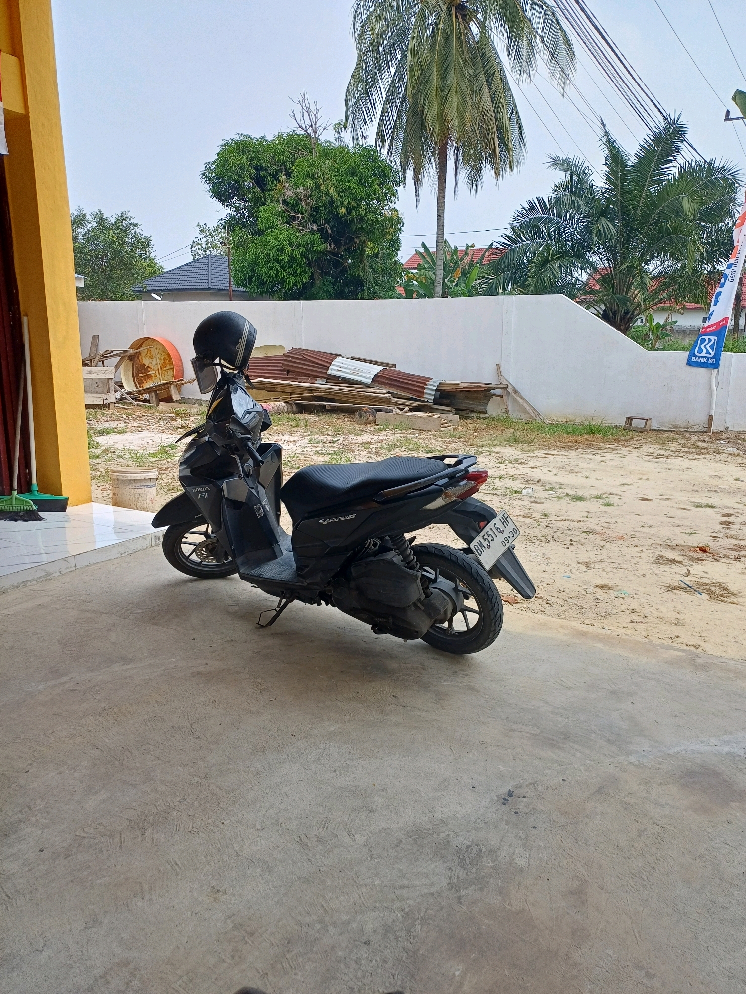 A black Honda motorcycle with a black helmet on the handlebars is parked on a concrete surface. The motorcycle has a license plate that reads "BM 5516 HF 09-20". In the background, there is a yellow building on the left, a white wall, scattered construction materials including corrugated metal sheets, and various types of trees, including palm trees. A flag with the "BANK BRI" logo is visible on the right side of the white wall. The ground appears to be a mix of sand and dirt. The sky is overcast. The scene is outdoors, likely in a residential or commercial area in Dumai City, Indonesia.