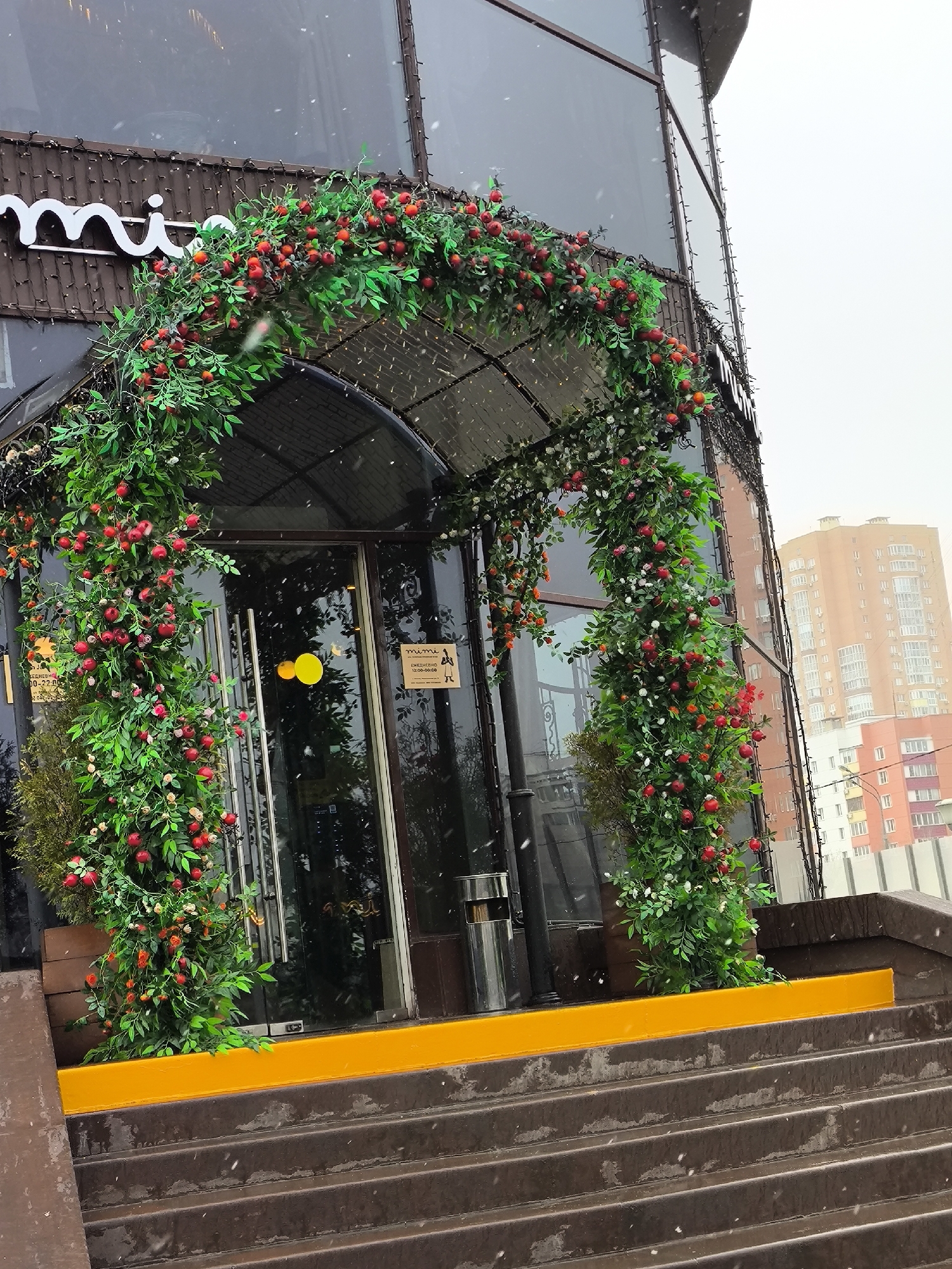 The image shows the entrance to a building, likely a commercial establishment, located in an urban environment. The entrance features a glass door adorned with an archway made of lush greenery interspersed with red and white flowers, possibly artificial. Two round, yellow stickers are placed on the door's glass panels. A sign in Russian is affixed to the glass, providing information such as business hours.
The entrance is accessed by a set of concrete steps with a yellow safety strip highlighting the edge. It's snowing lightly, as evidenced by the visible snowflakes. A tall building is visible in the background.