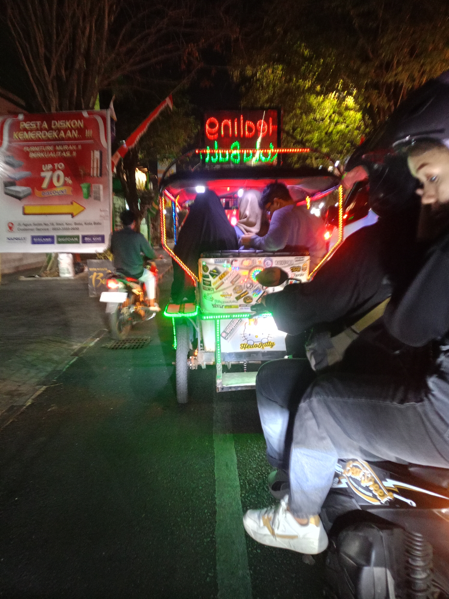 The image depicts a nighttime street scene in Batu, Indonesia. In the foreground, a person is riding on the back of a motorcycle, wearing a black jacket and light blue jeans. Their white sneakers with gold accents are visible. To their left, the side of a motorcycle is visible, featuring a gold and black design.
In the midground, a brightly lit motorized tricycle, known locally as a "Bentor," is moving away from the camera. The Bentor is decorated with numerous colorful lights and stickers, including a prominent "Hello Kitty" sticker on the side. Three individuals are visible within the Bentor: two passengers in the back, one wearing a dark hijab and the other a light-colored garment, and a driver in the front wearing a green long-sleeved shirt and white pants.
Behind the Bentor, a large billboard is illuminated, advertising a "Pesta Diskon Kemerdekaan" (Independence Day Discount Festival) with furniture offers and up to 70% discount. The billboard also shows product images of mattresses and furniture. In the background, trees with foliage are visible against the dark night sky, and red and white Indonesian flags are strung across the street. The street surface is dark asphalt, with a distinct green painted lane marker visible on the right side of the image. The overall atmosphere is lively and commercial, typical of a bustling street in an Indonesian city at night.