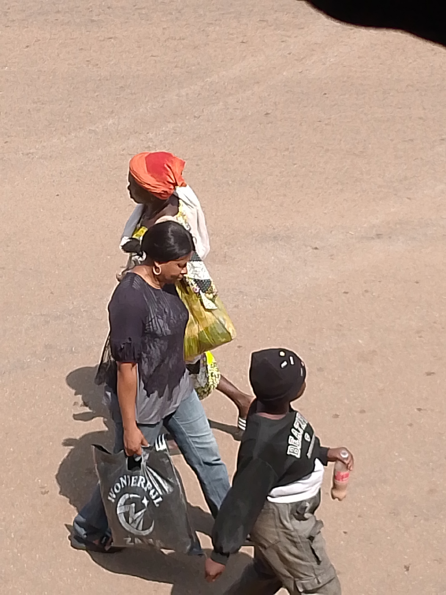 The image shows three people walking on a dusty, light-brown surface, likely a road or path. The perspective is from above, looking down on the subjects.
The first person from the background is wearing an orange headscarf and a white garment with a patterned trim. They are carrying a bright yellow bag.
In the middle ground, a woman with dark hair and large hoop earrings is walking. She is wearing a dark, possibly black, short-sleeved shirt and blue jeans. She is carrying a large black plastic bag with white text that appears to read "WONDERFUL" and a circular logo.
The third person, in the foreground, is a child wearing a black baseball cap and a black sweatshirt with white lettering on the back. They are also wearing grey cargo pants and holding a bottle of a brown beverage, possibly soda. The child's back is to the camera.
The scene appears to be outdoors, with shadows cast on the ground, suggesting it is daytime, possibly late morning or early afternoon, given the angle of the shadows. The weather seems dry. There are no other visible elements to indicate the specific location. The people appear to be walking with a purpose, possibly returning from or going to a market.