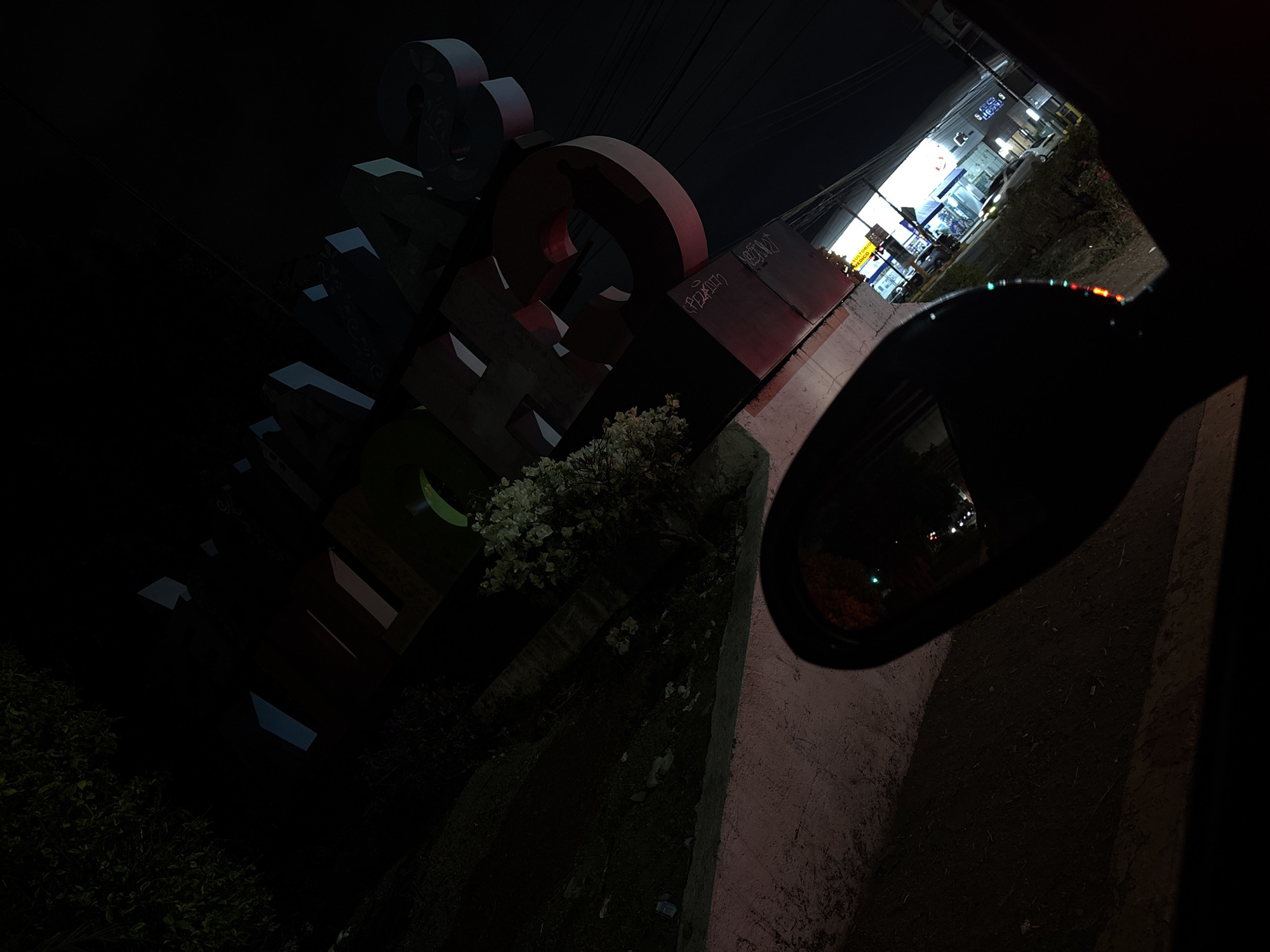 The image, taken at night, depicts a street scene in San Andrés Cholula, Mexico. A large, illuminated sign with blocky, three-dimensional letters, primarily red, dominates the left side of the frame. The letters are partially obscured by a flowering plant. A sidewalk runs diagonally from the bottom right towards the center, with a car's side mirror visible in the foreground. Across the street, a brightly lit convenience store is visible, displaying signs in Spanish. Cars are parked outside the store. Overhead wires crisscross the sky. The overall scene is dark, with lighting primarily provided by the store and the illuminated sign.