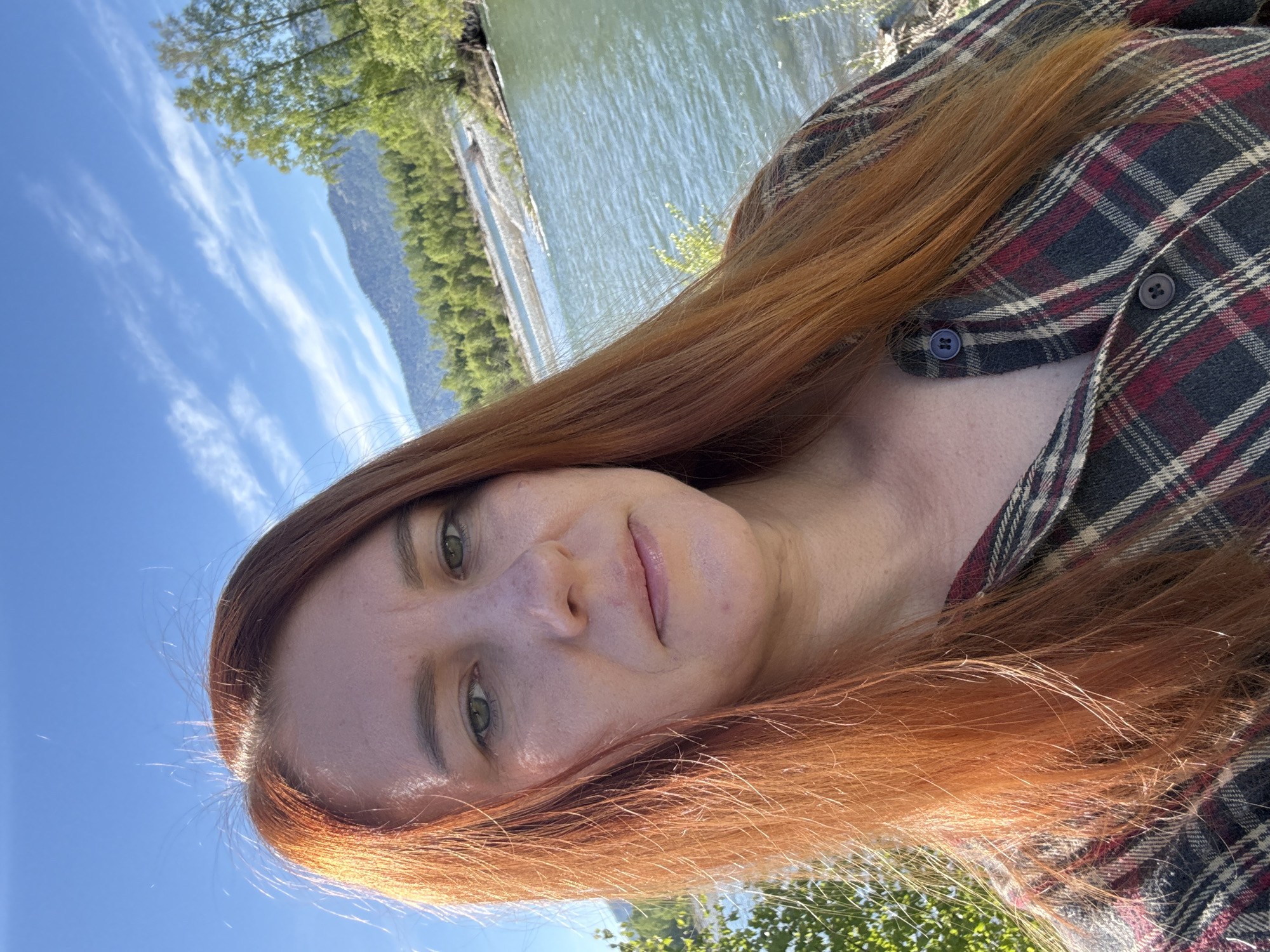 A woman with red hair is pictured in an outdoor setting. She is wearing a plaid shirt. Behind her is a body of water and a treeline. The sky is blue with streaky clouds. The location is likely Ust-Koksa, Russia, given the prompt, but there is no way to confirm this from the image alone.
