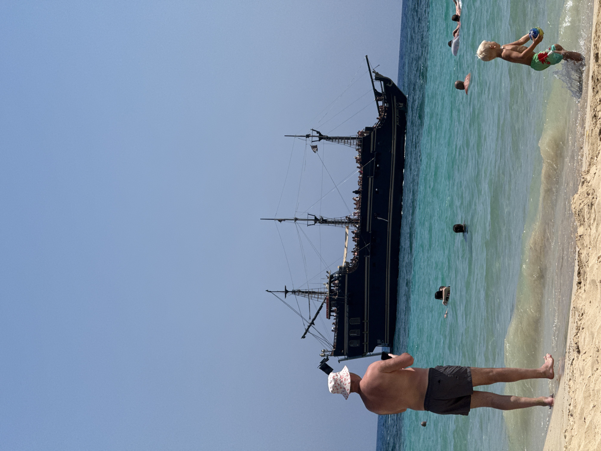 A man wearing a bucket hat and swim shorts stands on a sandy beach in Ayia Napa, Cyprus, holding a phone. In the background, a large, dark-hulled pirate ship with multiple masts sails on the turquoise sea. Several people are visible in the water, some swimming, and others relaxing. A young boy in green swim trunks plays with a ball near the water's edge. The sky is clear and blue.