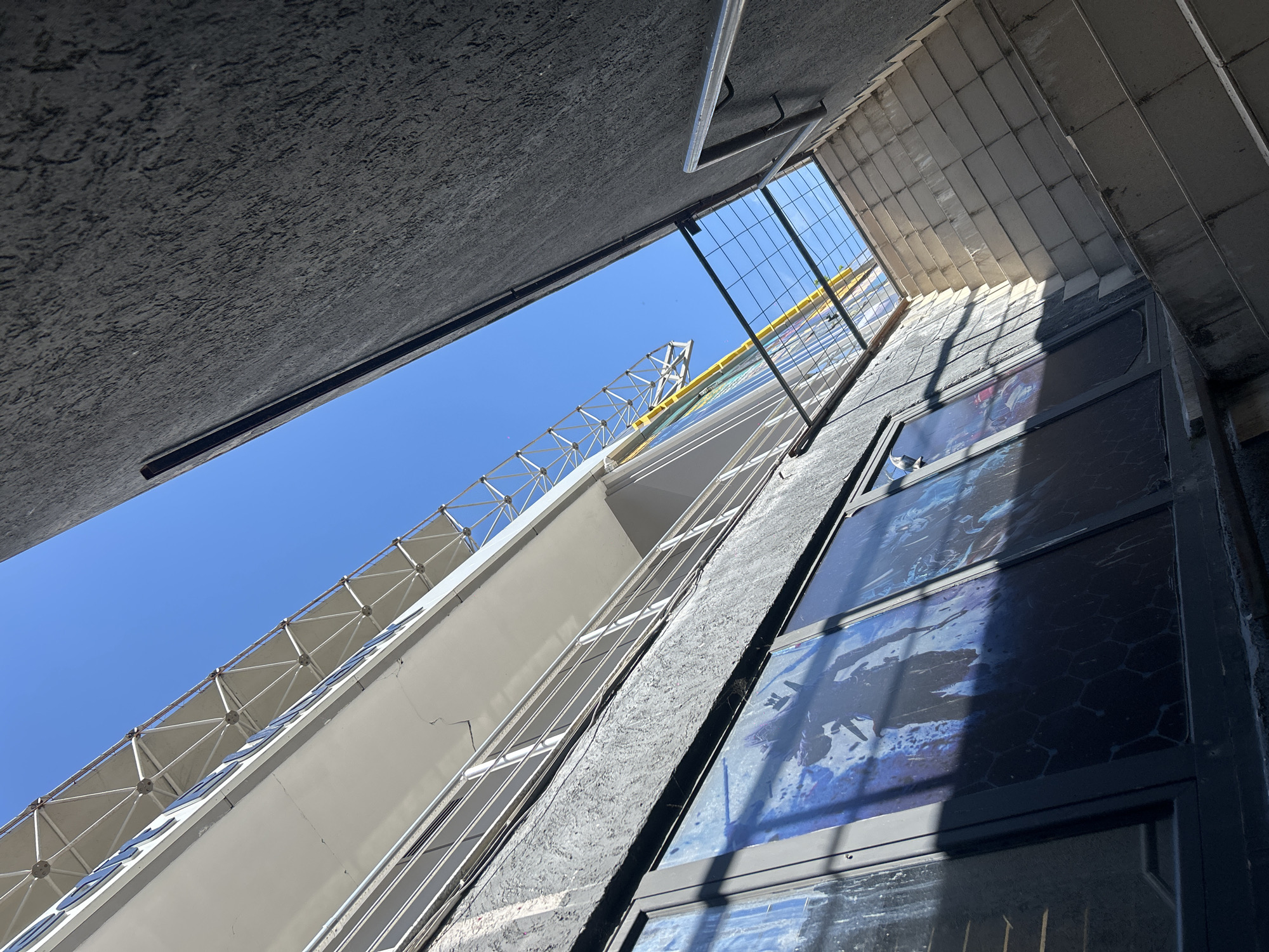 The media file is a low-angle, upward-looking shot of a modern building with a clear blue sky visible. The architecture features geometric lines, with a predominantly white structure contrasting against textured gray surfaces. A metal railing runs along a staircase, and a wire mesh fence is also visible. The building has large windows with what appear to be artistic or abstract blue and white patterns within them, casting shadows on the surfaces. Sunlight is strong, suggesting a bright, clear day. The perspective creates a sense of height and depth, emphasizing the scale of the building. There are no people or explicit activities taking place. The overall impression is one of urban architecture under a sunny sky. The location context provided is Burgas, Bulgaria.