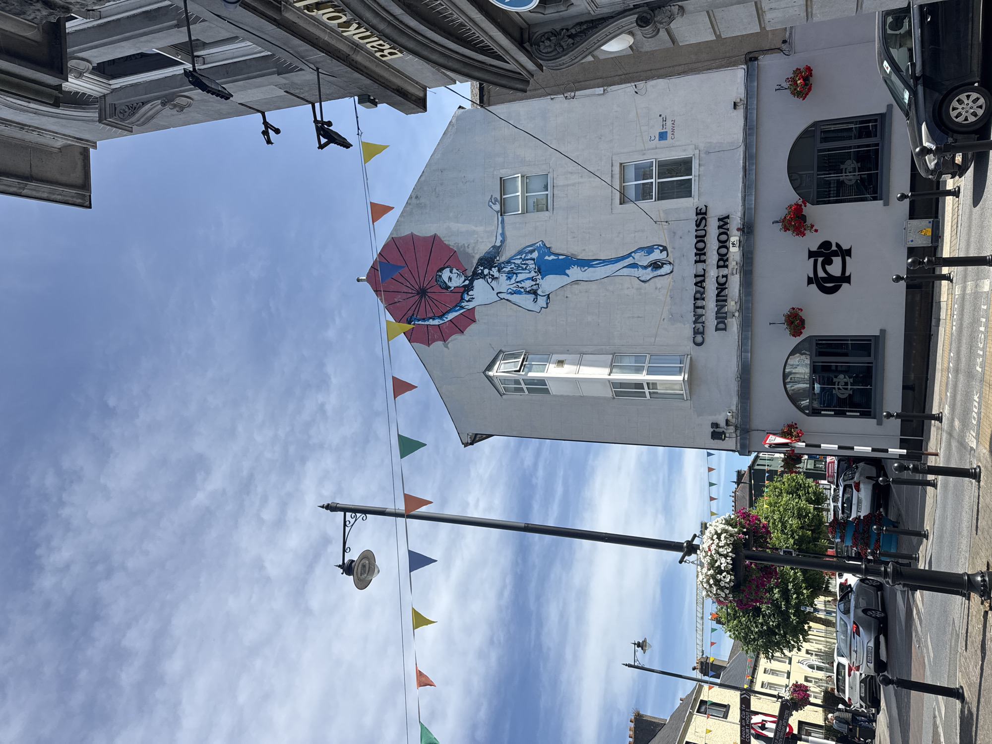 The image depicts a street scene in Clonmel, Ireland, on a sunny day with a partly cloudy sky. A large mural of a ballet dancer holding a red umbrella adorns the side of a building labeled "CENTRAL HOUSE DINING ROOM." The building is light gray and has several windows. To the right, a section of a building with the words "Bank of I" visible suggests the presence of a bank. Colorful triangular flags are strung across the street between lampposts. A lamppost with a hanging basket of flowers stands in the foreground. Several cars are parked and driving on the street, which is paved with asphalt. Pedestrians are visible in the background, and signage indicates that this is a town center. The overall impression is a vibrant urban environment with a touch of artistic flair.