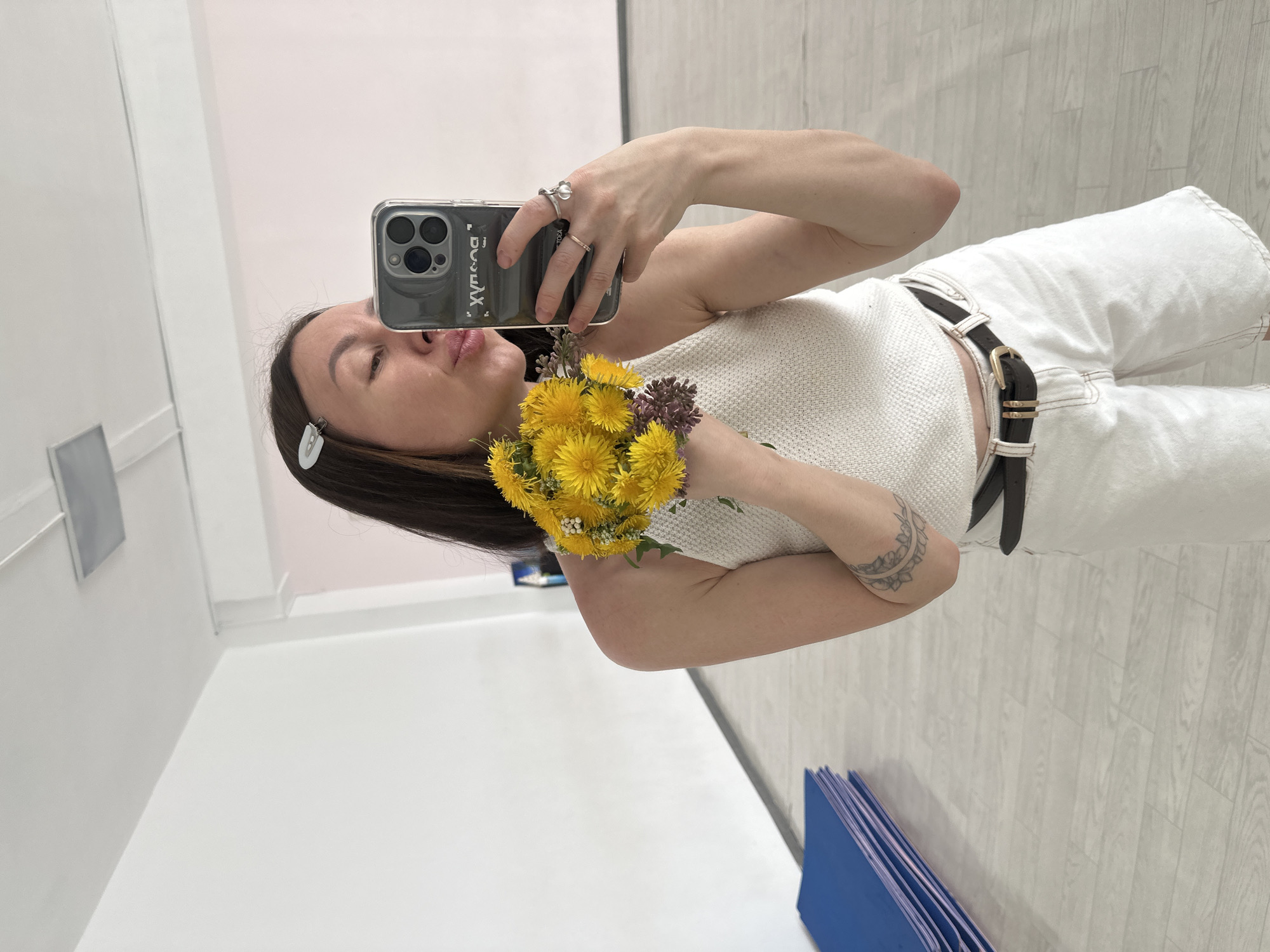 A woman stands in a room, taking a selfie with her smartphone. She holds a bouquet of dandelions and purple wildflowers in front of her. She is wearing a white knitted top, white jeans with a black belt, and has a hair clip in her dark hair. The room has white walls and a light-colored wood-patterned floor. Blue foam mats are stacked in the corner. The reflection in the phone's screen suggests she is in a room with light pink accents near the ceiling and a possibly a mirror. There is no clear indication the location is Votkinsk, Russia, or any other specific location.