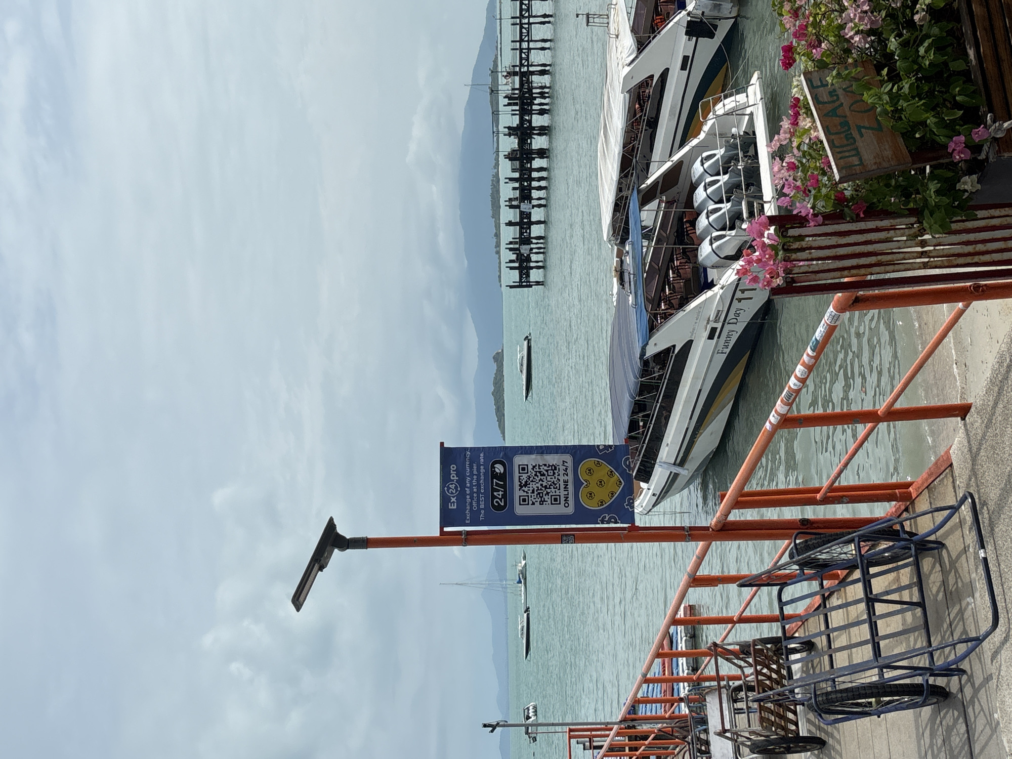 The image shows a coastal scene in Baan Bang Rak, Thailand. The sea is visible with several boats docked and anchored. A long pier stretches out into the water. The sky is overcast with a light gray tone.
In the foreground, there is a sign on a pole advertising "Ex24.pro" for currency exchange, emphasizing "24/7" availability with a QR code. Next to the sign, there's a boat named "Funny Day 11" docked at the pier. A wooden sign with "LUGGAGE ZONE" is also visible amidst green foliage and pink flowers. Luggage carts are parked along the pier edge, held back by orange guardrails.
