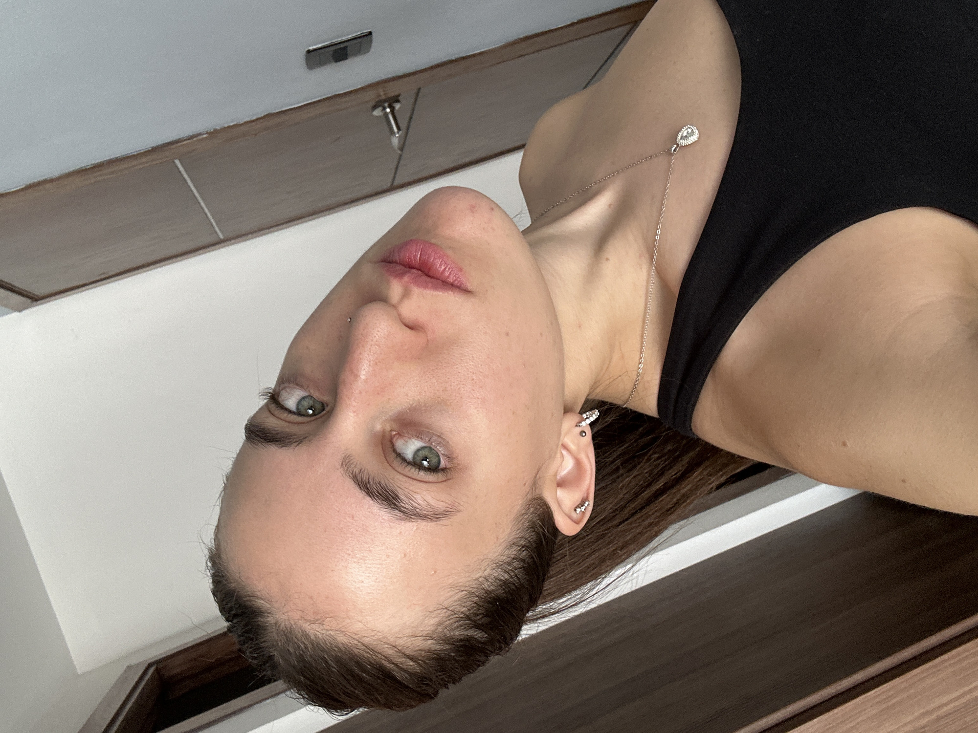 A selfie of a fair-skinned woman with green eyes and brown hair. She is wearing a black tank top and a delicate gold necklace with a teardrop-shaped pendant. She has multiple piercings in her left ear and a piercing above her lip on the left side of her face. The background shows a wooden cabinet, a white wall, and a light fixture. The location of Huixquilucan de Degollado, Mexico, cannot be confirmed based on the visual information.