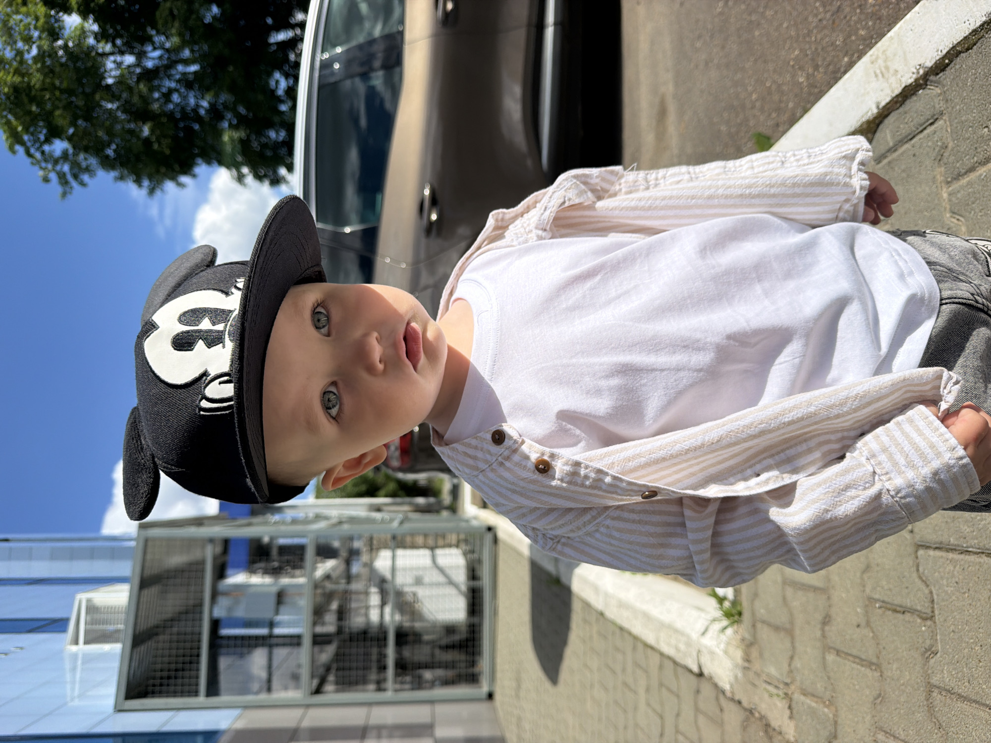 A young child wearing a white t-shirt, a striped unbuttoned shirt, and a black Mickey Mouse baseball cap stands outdoors on a paved surface, likely a sidewalk, in Chaikovsky, Russia. The child's eyes are a striking blue, and their lips are slightly parted. Behind the child, a modern building with glass and metal elements is visible, along with a large green tree and a metallic brown car parked on the street. The sky is a clear blue with some scattered white clouds. The lighting suggests it is daytime, with shadows cast by the child and the building. The overall scene depicts a casual outdoor moment in an urban environment.