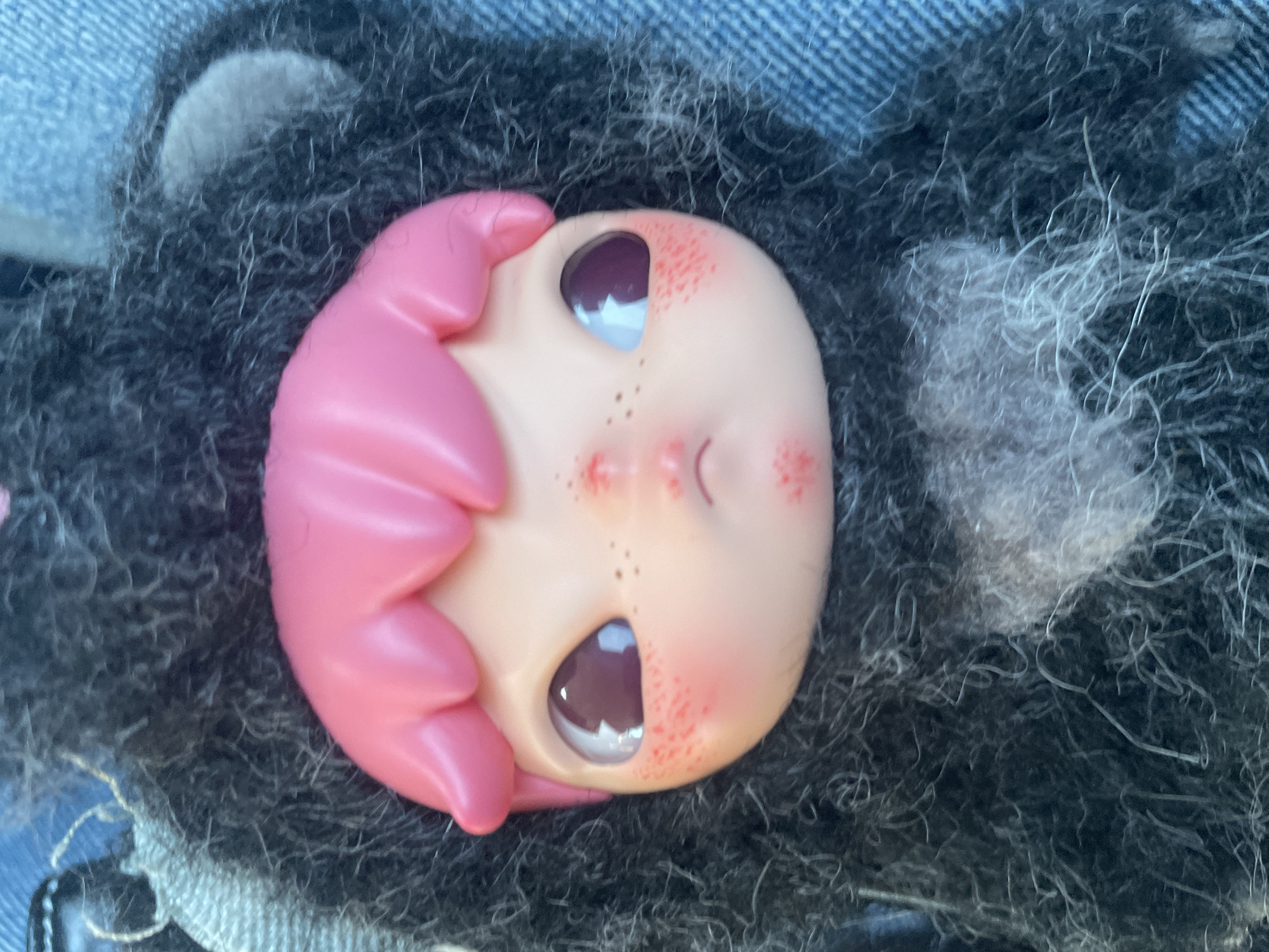 A close-up shot depicts the face of a doll with pink hair lying on a dark, furry surface, partially resting on denim fabric. The doll's face is fair with freckles and large, dark eyes. No information in the image suggests a location in Denpasar, Indonesia.