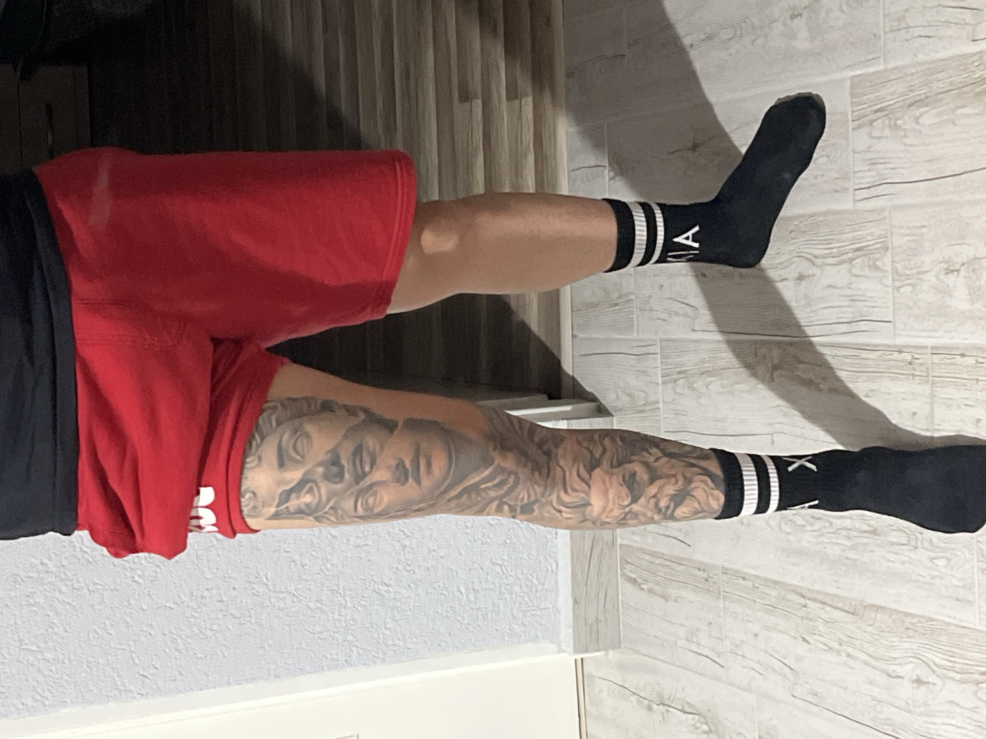 The image shows a person wearing red shorts, a black shirt, and black socks with white stripes. The person has a tattoo on their left leg. The background appears to be a room with wood-patterned flooring and a textured wall. There is no information to ascertain the location of the photo in Elhovo, Bulgaria.