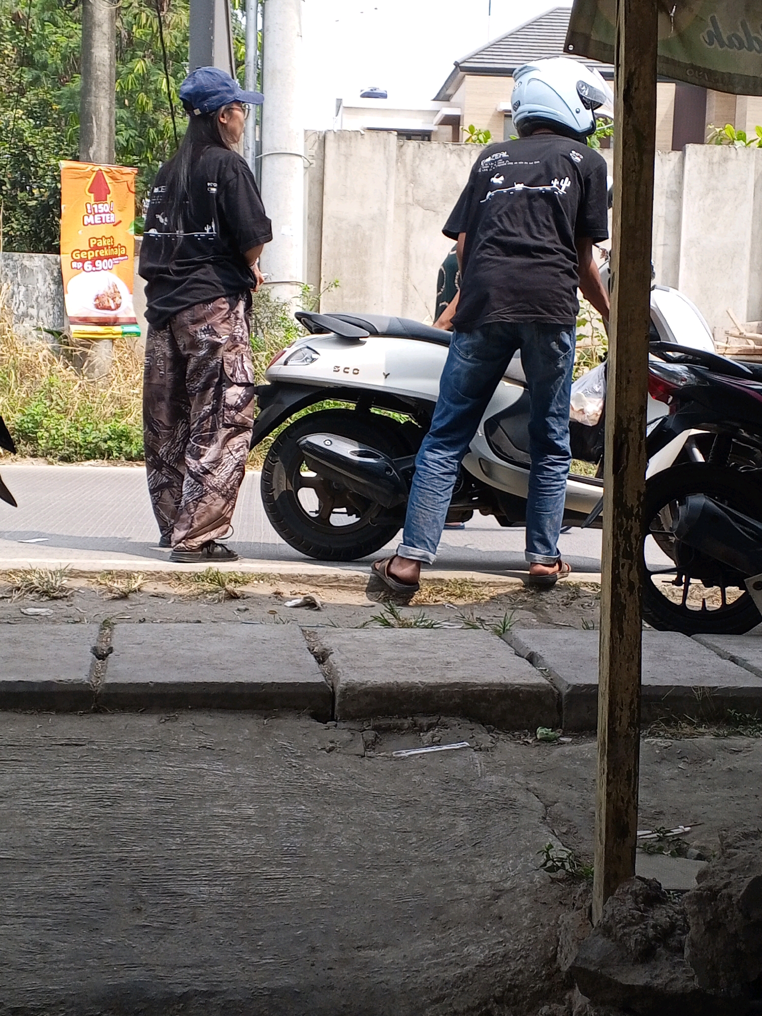 In the city of Ranjeng, Indonesia, two individuals stand near a scooter on a paved road. The person on the left, facing away from the camera, wears a black t-shirt, a blue cap, glasses, and camouflage pants. The person on the right, also facing away, is wearing a black t-shirt and blue jeans, with a light blue helmet on their head. The scooter is white and silver, parked on the side of the road. A street sign is visible in the background, along with a concrete wall and some vegetation. The scene appears to be outdoors during the daytime, with natural lighting.
