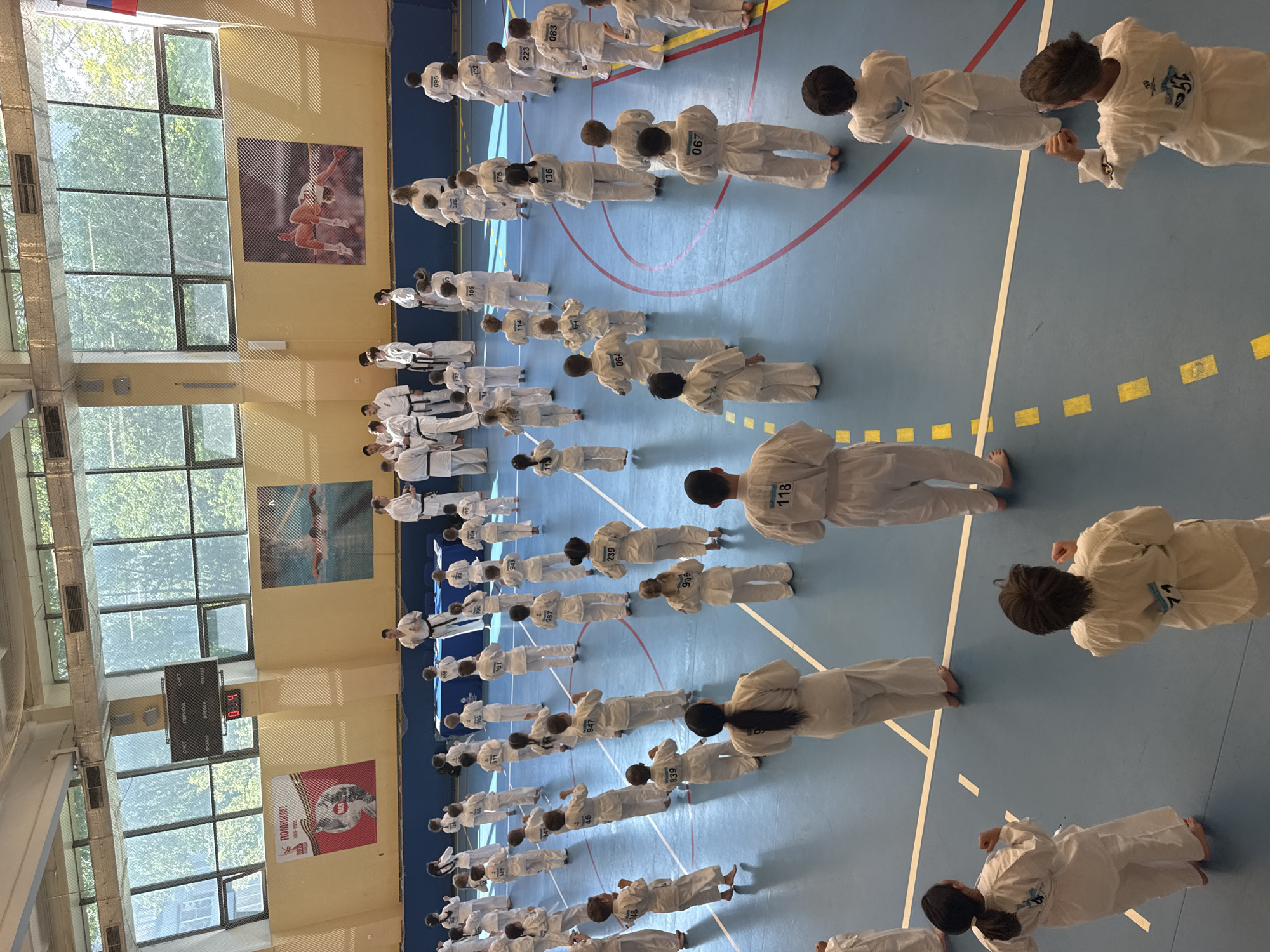 The image depicts a large group of people, predominantly children, in a gymnasium. They are arranged in rows, all wearing white martial arts uniforms. Numbers are affixed to their backs. A few adults in similar uniforms but with black belts are visible at the front of the formation. They seem to be instructing the group. The gymnasium has a blue floor with various lines and markings. Large windows line one wall, providing natural light and a view of trees outside. Posters of athletes are displayed on the walls between the windows. An electronic scoreboard is also visible. The location is in the city of Yekaterinburg, Russia. The overall scene suggests a martial arts class or training session in progress.