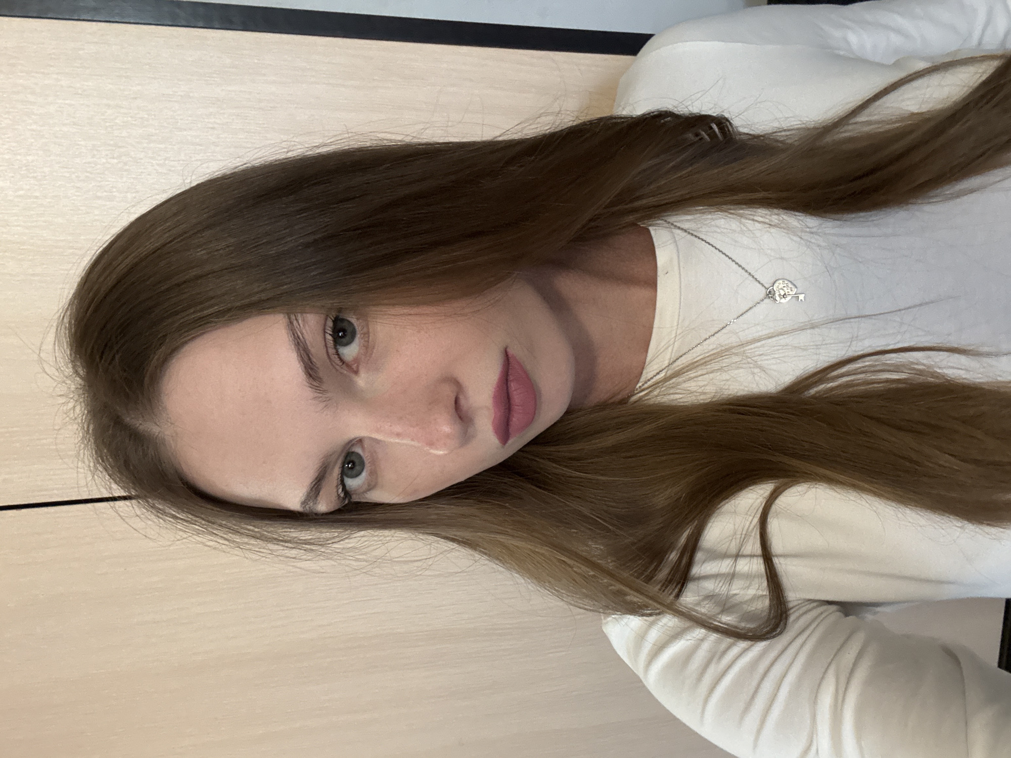A young woman with long brown hair, wearing a white long-sleeved shirt and a silver necklace with a heart and key pendant, is pictured in a close-up selfie. She has light skin, green eyes, and is wearing matte pink lipstick. The background appears to be an interior space with a light-colored wooden wall and a dark vertical element on the right. There is no indication of a specific location within the city of Perm, Russia, or any actions or interactions taking place.