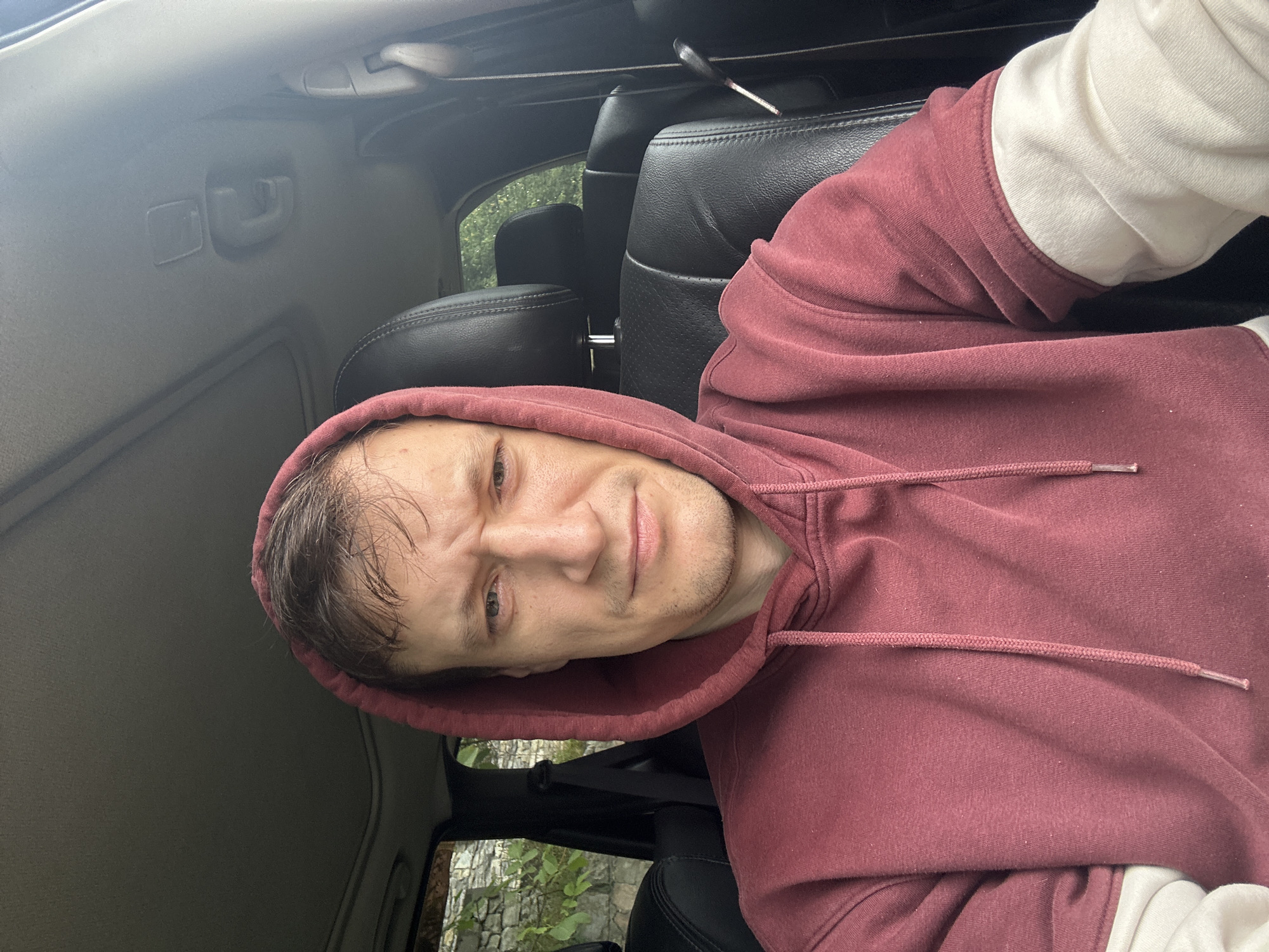 A man is sitting in the driver's seat of a car, looking at the camera. He is wearing a maroon hooded sweatshirt. The car's interior is visible, including the dashboard, headrests, and a sunroof. Outside the car, foliage and a stone wall can be seen through the windows. The scene appears to be an outdoor location in Makiv, Ukraine.