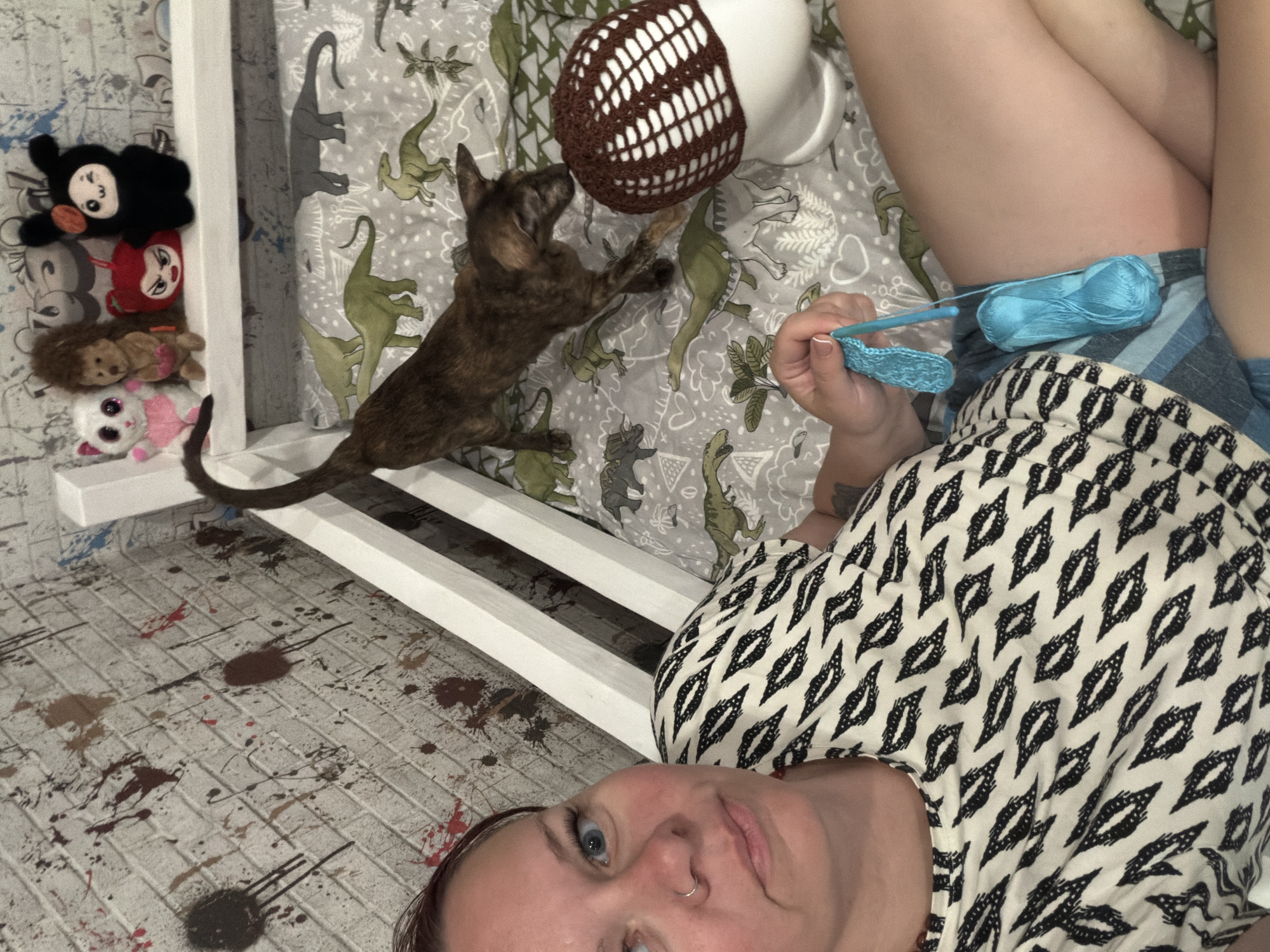 A woman with blue eyes and a nose ring is taking a selfie while lying on a white-framed bed. She is wearing a black and white patterned top and blue shorts. She is holding a blue crochet hook with blue yarn, creating a crochet piece. A brindle-colored dog is on the bed, appearing to interact with a white mug topped with a brown and white crocheted object. The bed has dinosaur-themed bedding. To the left of the bed, there are several stuffed animals. The floor appears to have paint splatters. The location cannot be determined, as the prompt is outside the scope of the available information.